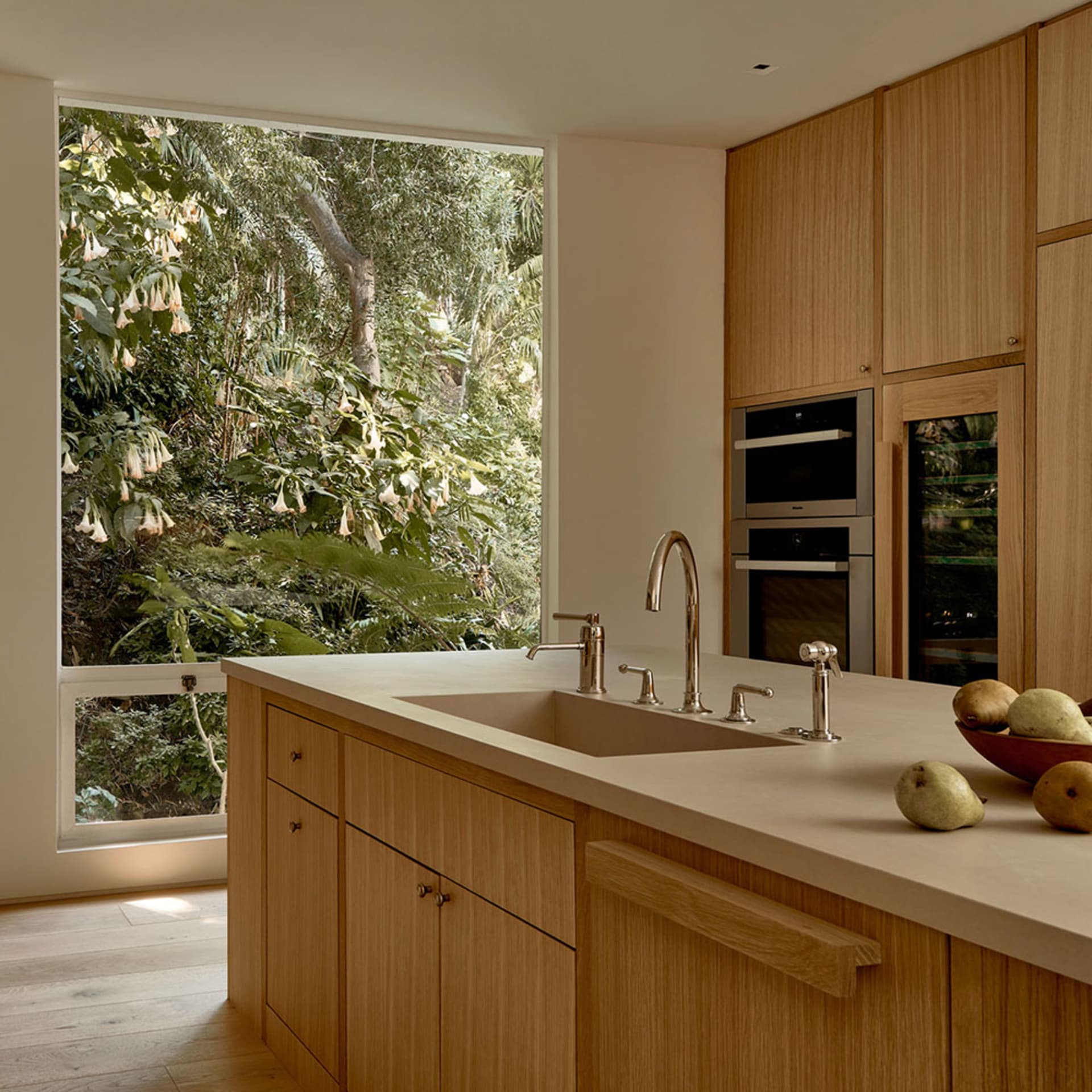 A modern kitchen with wooden cabinets, a sink, and fruits on the countertop, overlooking a lush green outdoor scenery through