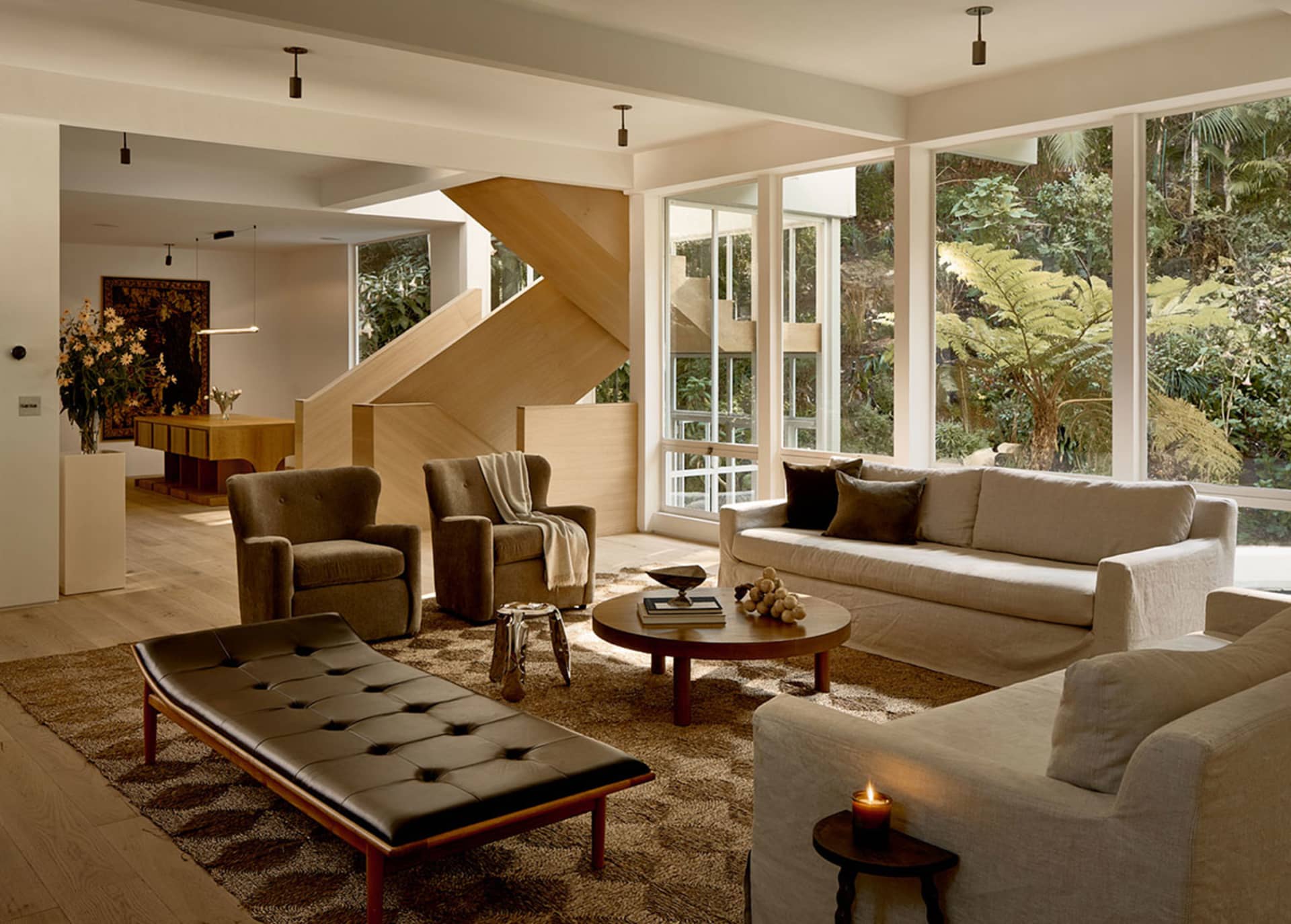 A modern living room with a wooden staircase, a leather ottoman, and a beige sofa with a round wooden coffee table.