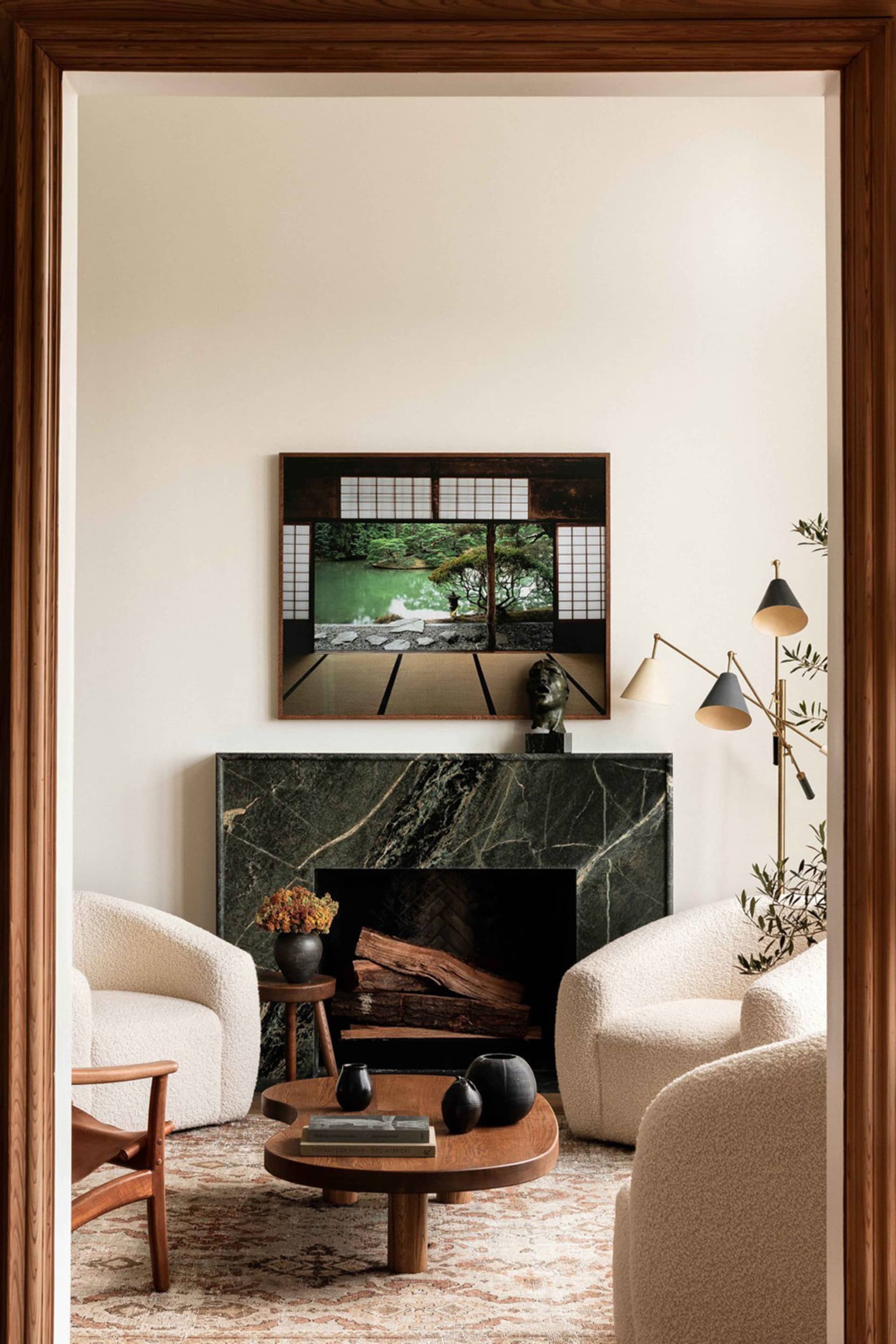 A modern living room with a marble fireplace, white upholstered chairs, a wooden coffee table, and an abstract painting.