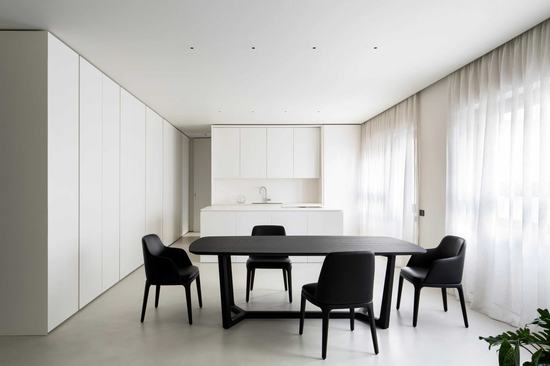 A modern minimalist kitchen and dining space with white cabinets, black furniture, and sheer curtains.