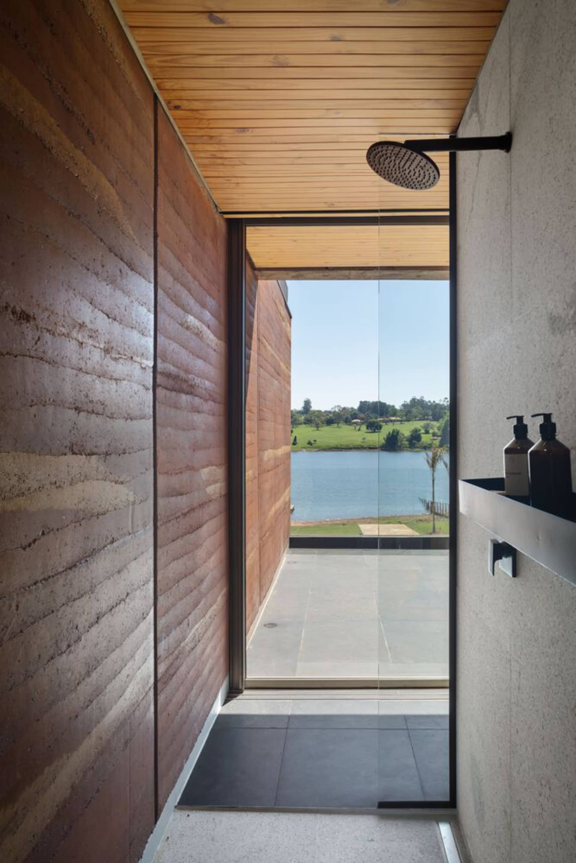An industrial-style bathroom with a wooden shower area and a view of a tranquil lake through a large glass window.