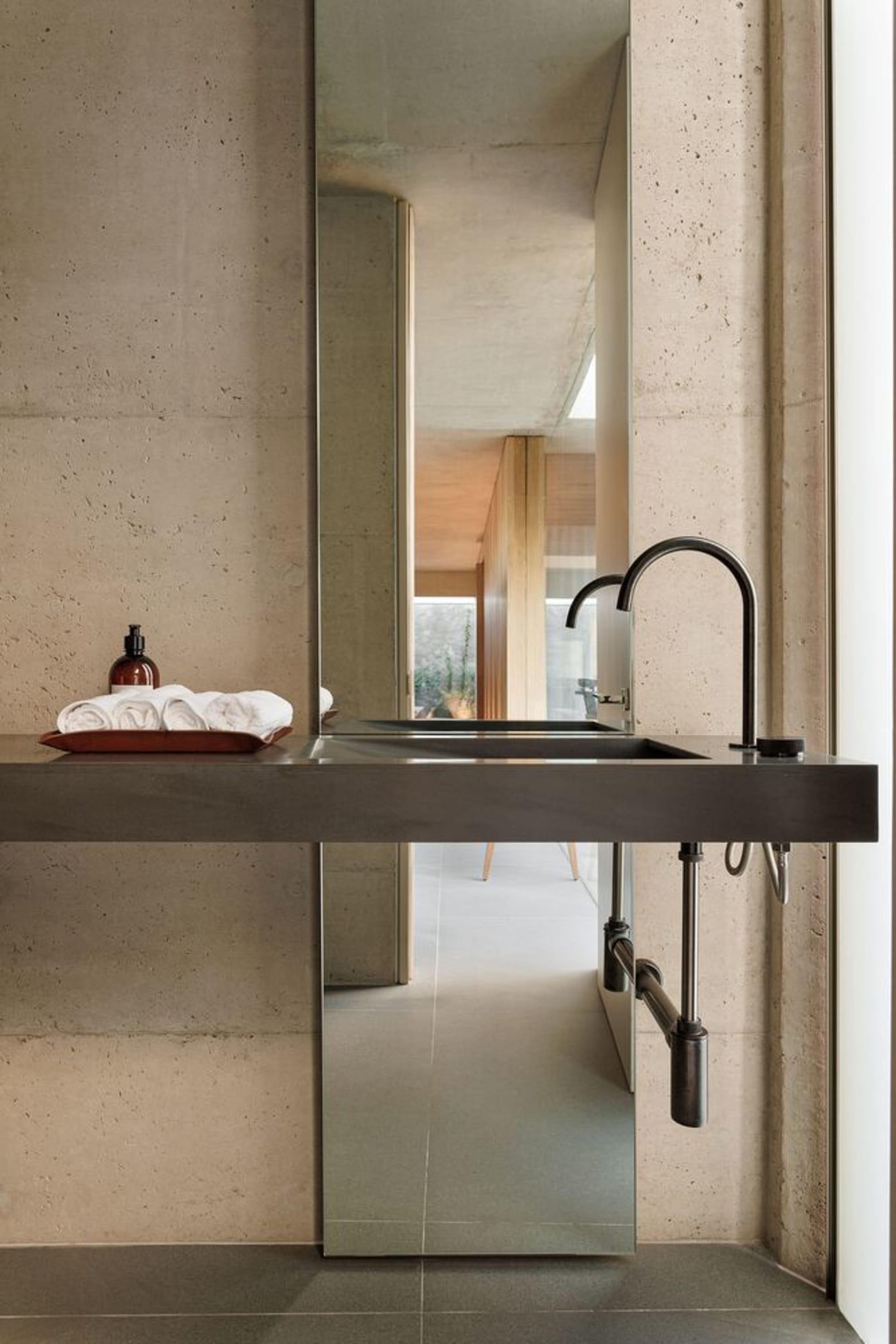 Modern minimalist bathroom with concrete walls and floor, featuring a long sink with a black faucet and a large mirror above.