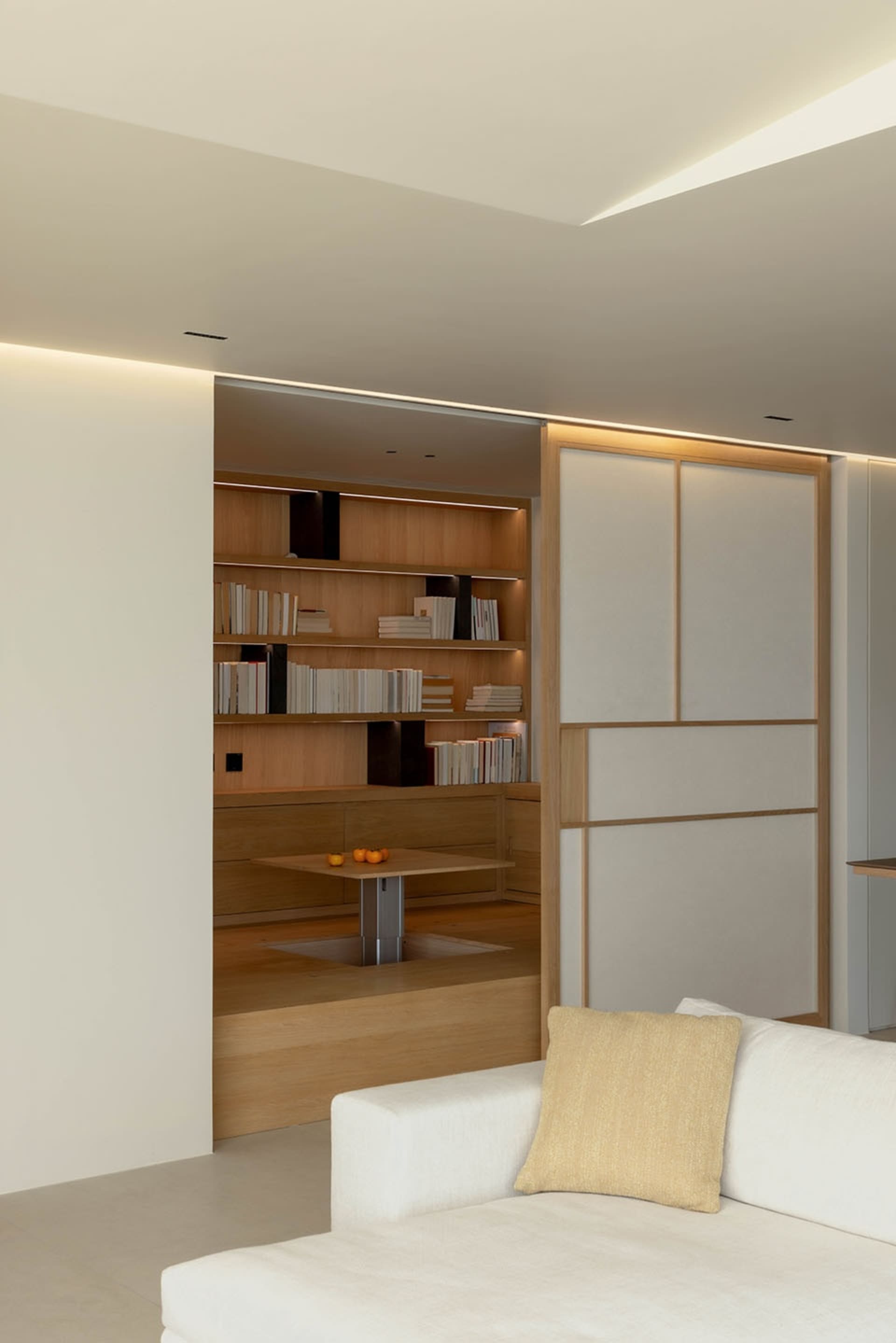 A modern minimalist living room featuring a white couch, a wood coffee table, and a bookshelf filled with books.