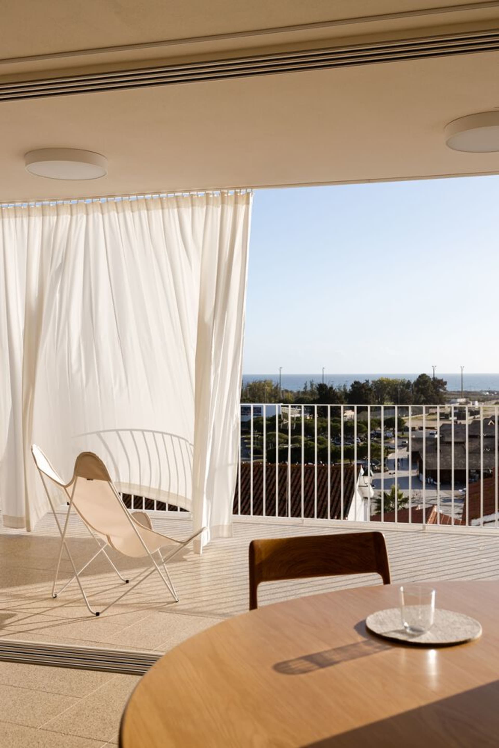 A minimalist residential patio features a beige and white upholstered chair, a round wooden table with a glass drink containe