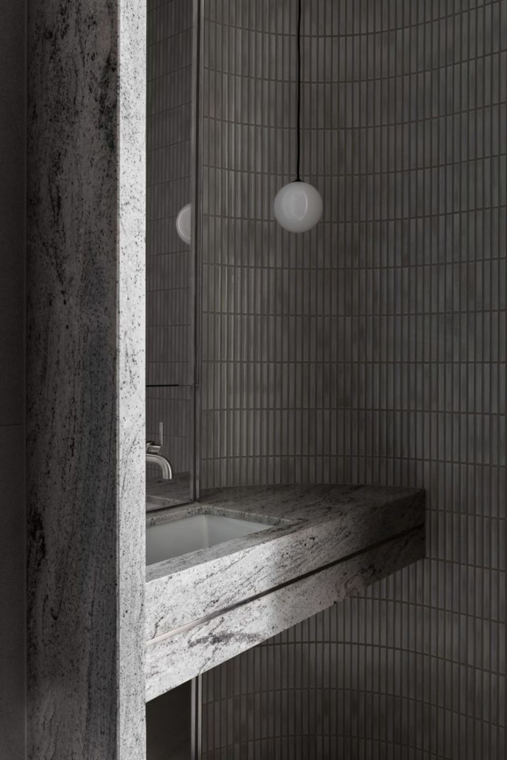 A minimalist bathroom with gray walls covered in small, vertical tiles and a granite countertop featuring a white sink.