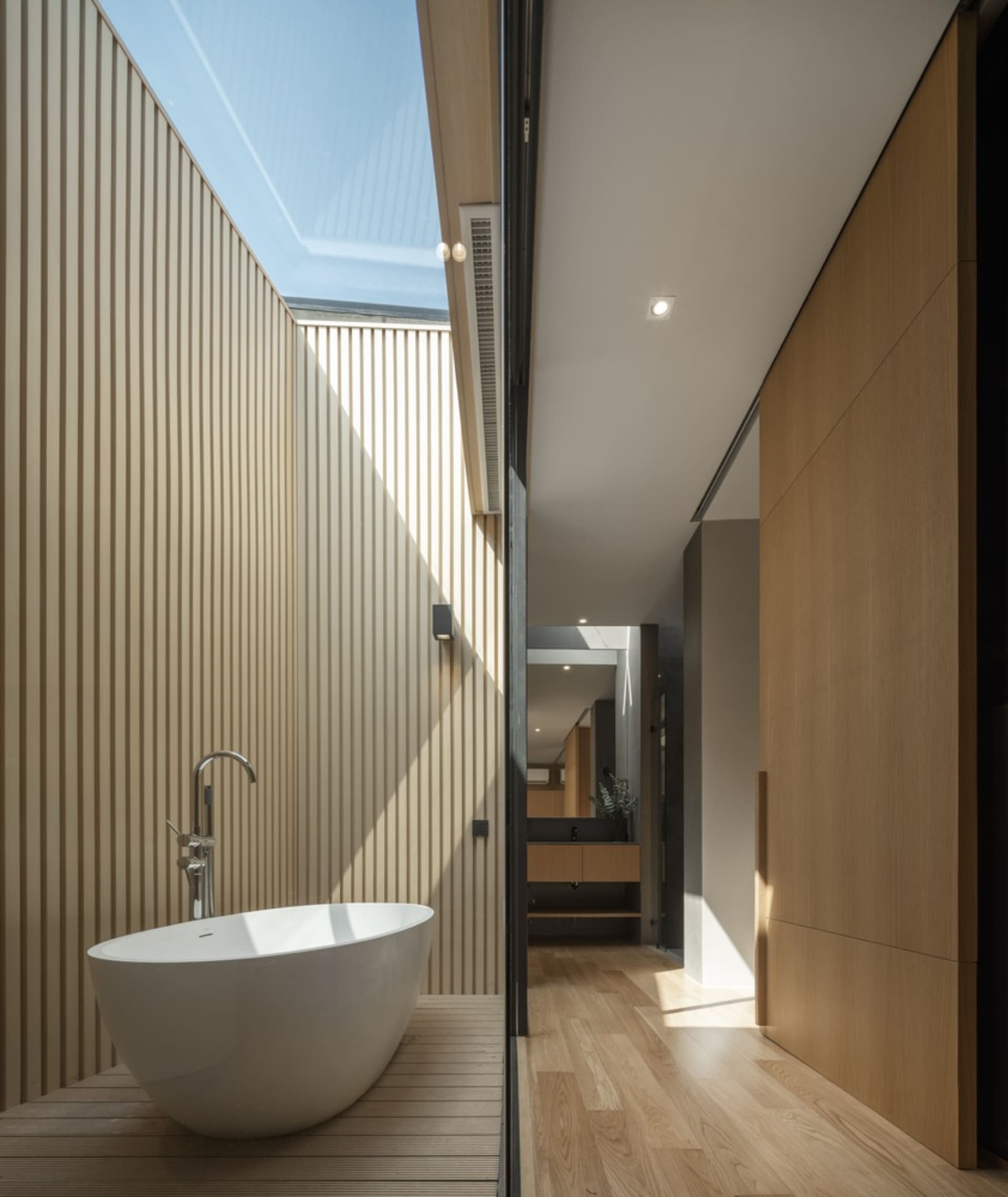 Modern minimalist bathroom with a white stand-alone tub set against a wooden floor and wall, featuring a skylight for natural