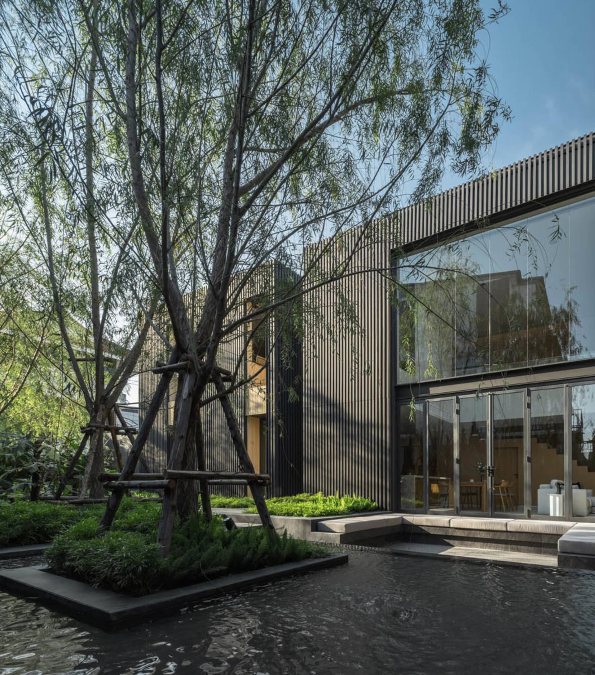 A modern residential exterior featuring a combination of wood and glass materials, set against a natural tree and water featu