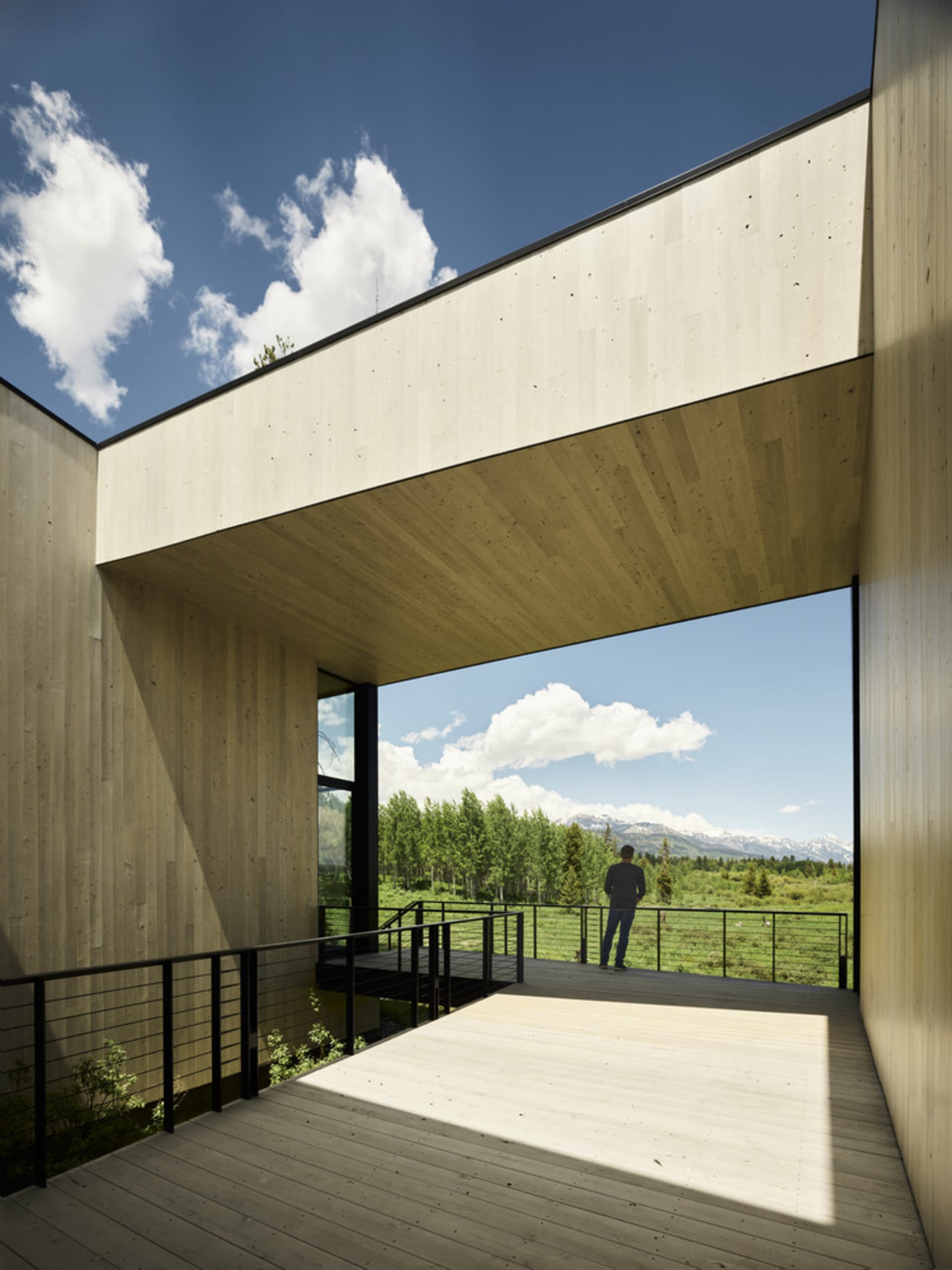 Modern exterior view of a building with wooden panels and concrete roof, featuring a person standing on a wooden deck overloo