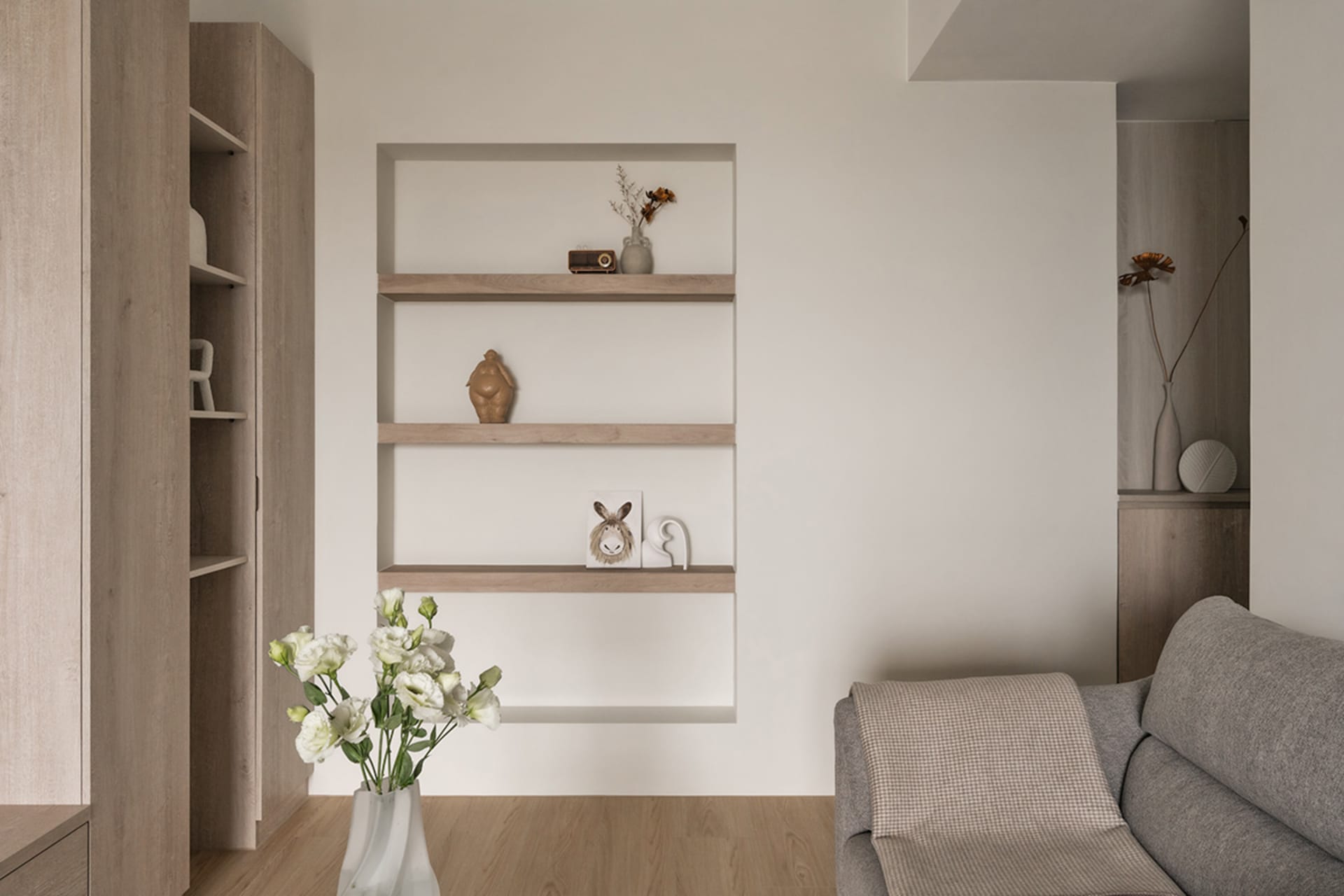 A minimalist living room featuring a gray couch, white flowers in a vase, and a floating shelf with decorative items on a neu