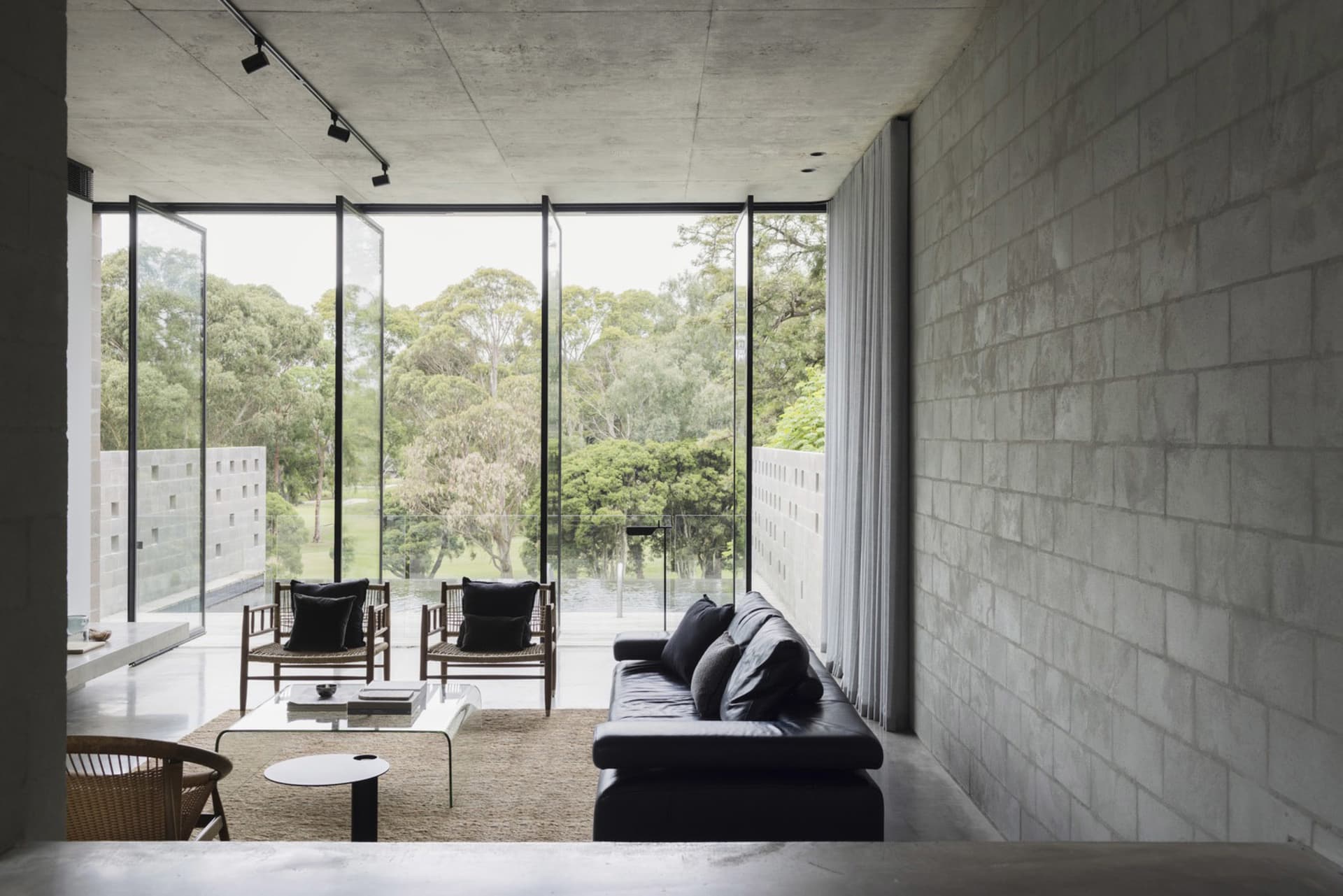 A modern living room with concrete walls, a black leather sofa, and large windows overlooking a green landscape.