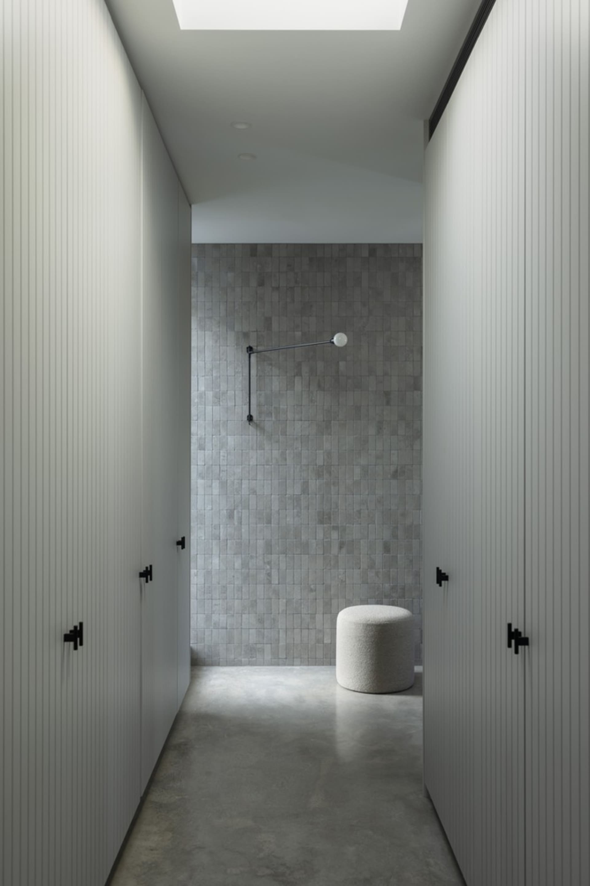 Modern minimalist bathroom with concrete tiles and floor, featuring a single lamp and a white stool.