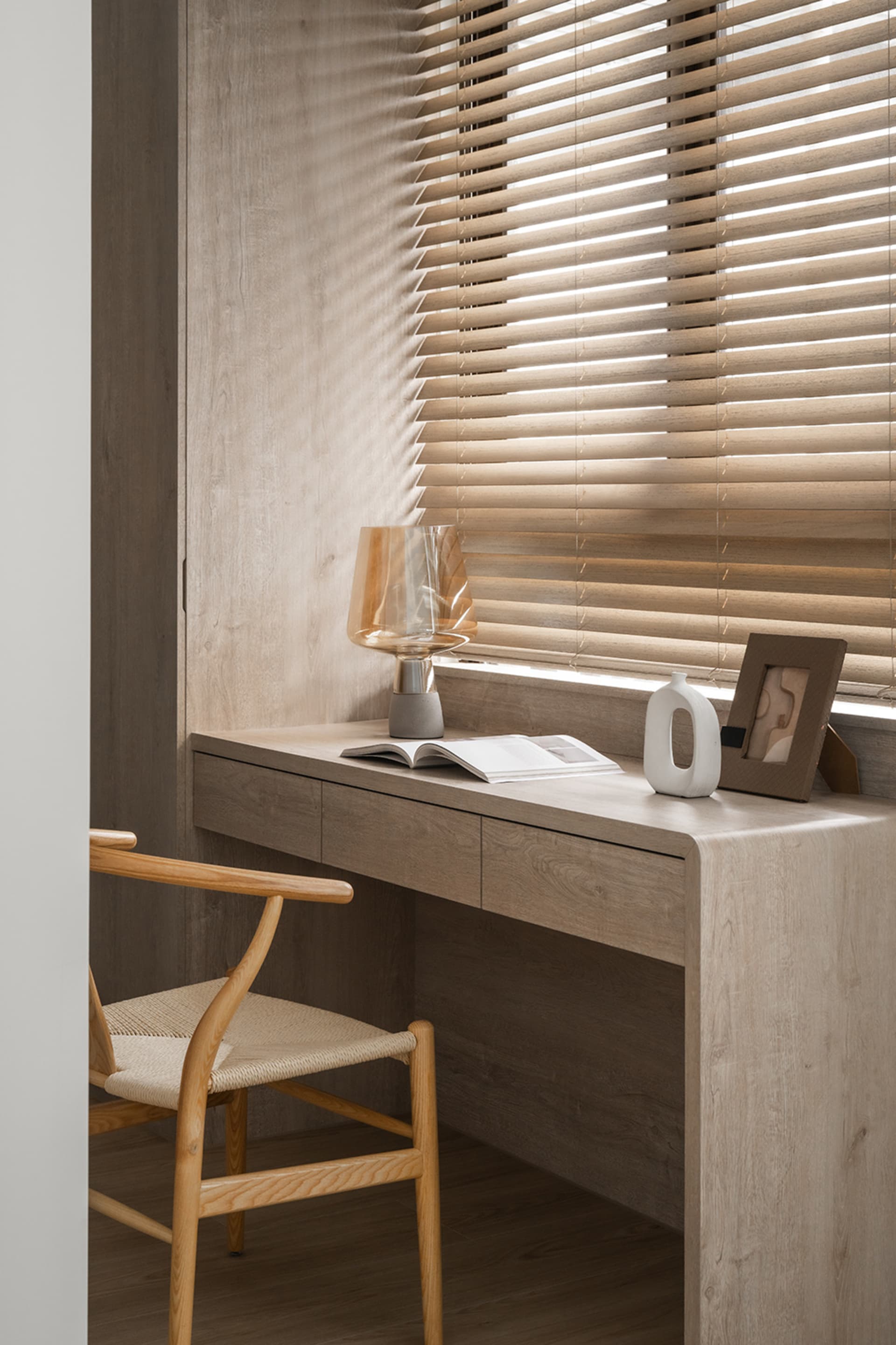 A minimalist study area featuring wooden furniture, a glass lamp, and a book with a vase and a framed picture on a wooden des
