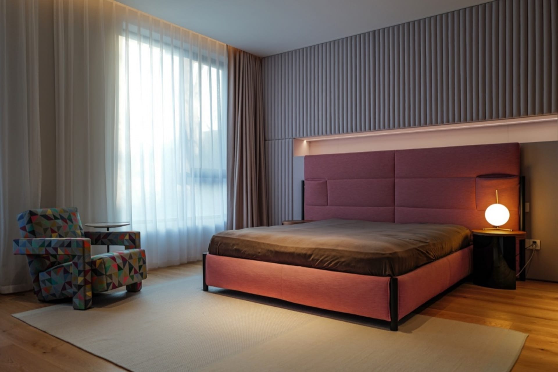 Interior design of a minimalist bedroom featuring a bed with a large burgundy headboard and an armchair with a geometric prin