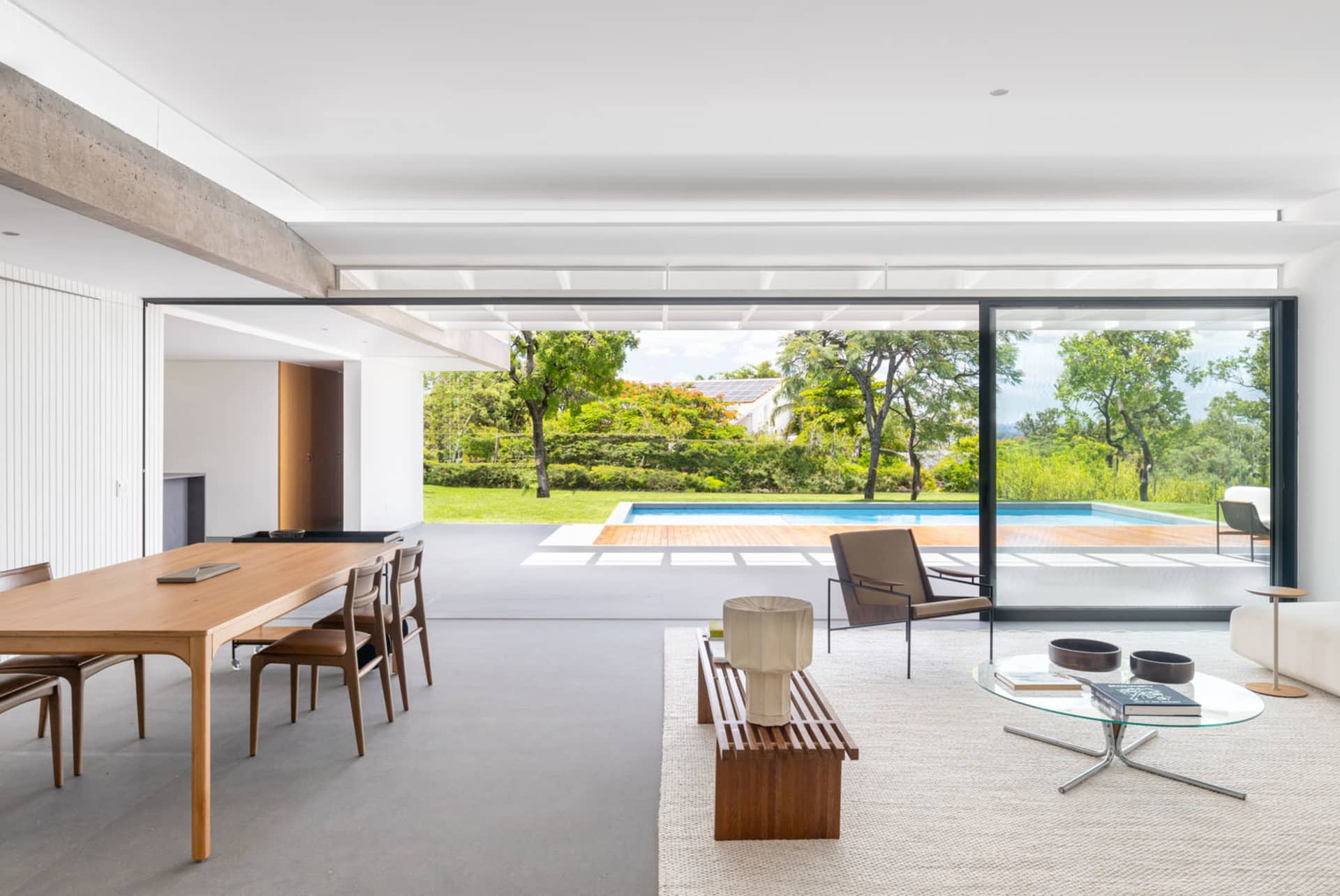 A modern living room featuring wooden tables, chairs, and a glass coffee table with a view of a pool and greenery.