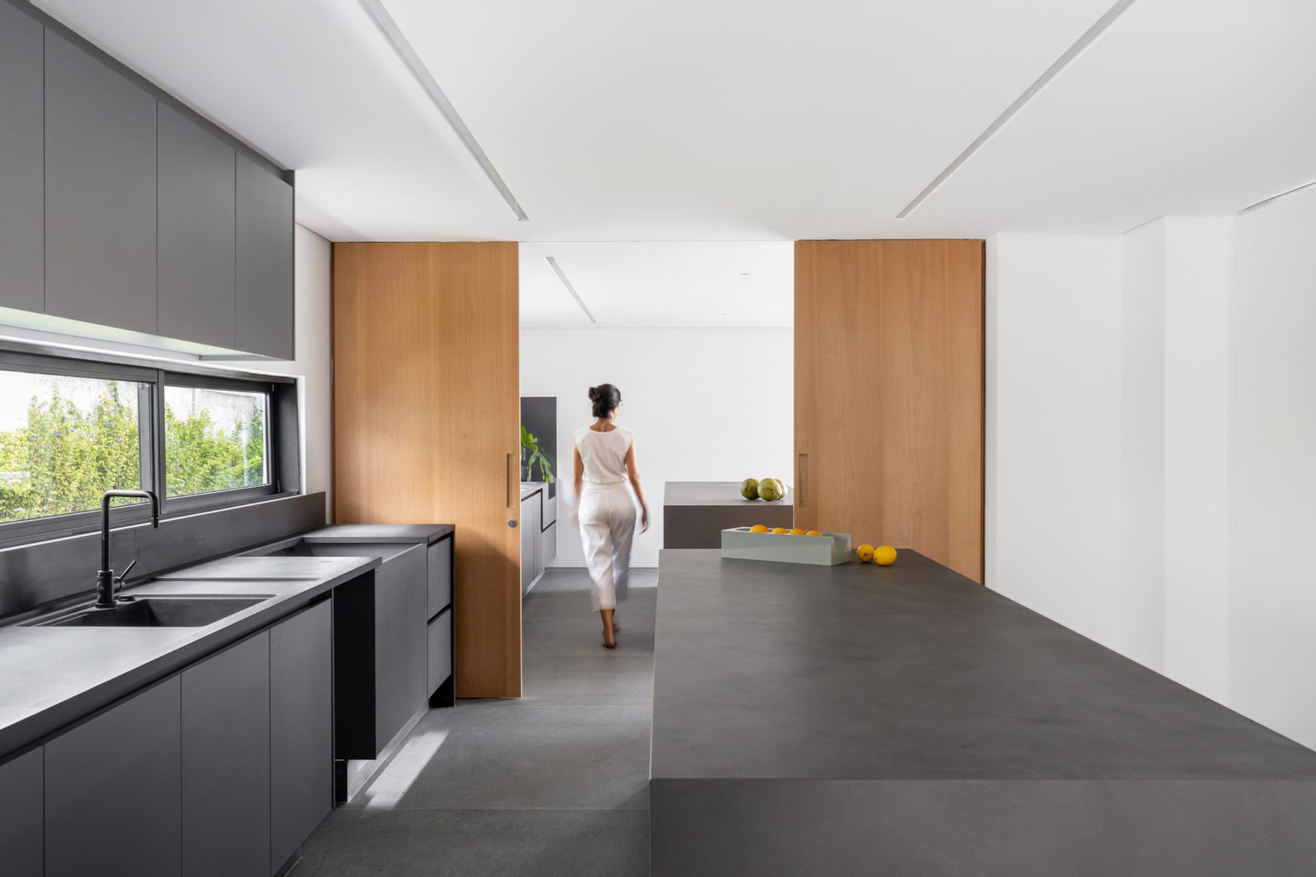 A modern minimalist kitchen with concrete countertops and wooden partitions, featuring a person in white attire.