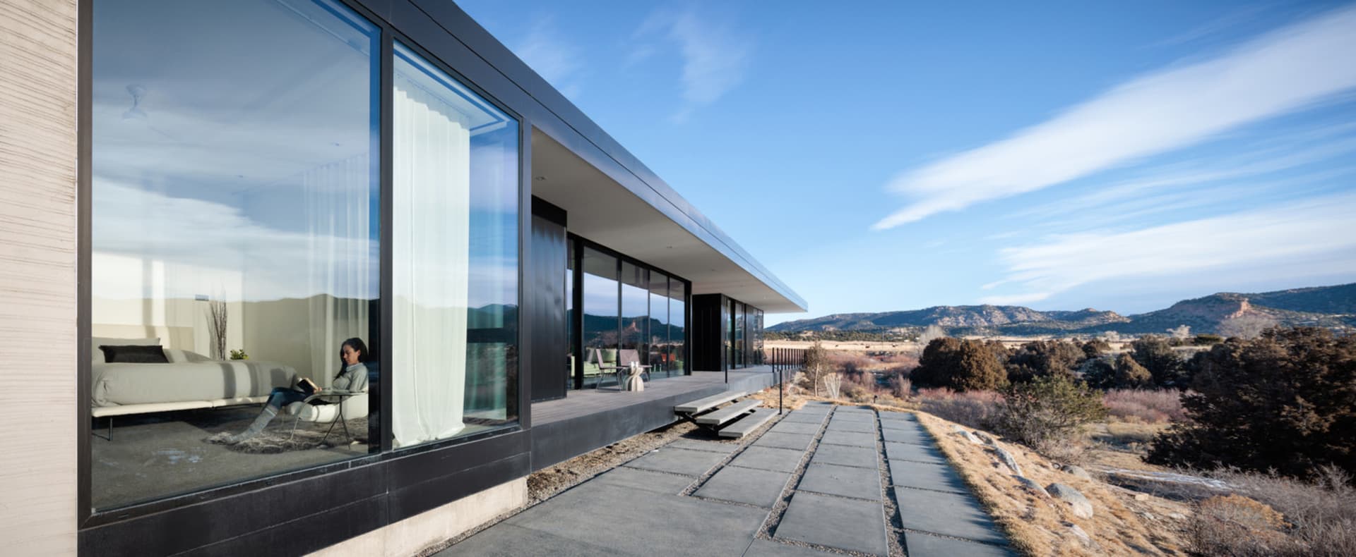 Modern minimalist residential exterior featuring glass walls, concrete patio, and steel accents against a blue sky.