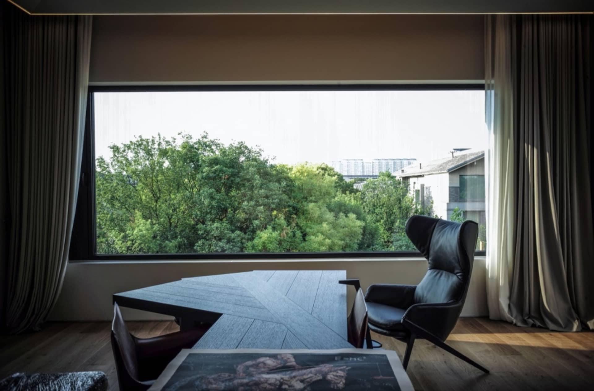 A modern office space featuring a large window with a view of lush greenery, a sleek black desk, and comfortable black chairs