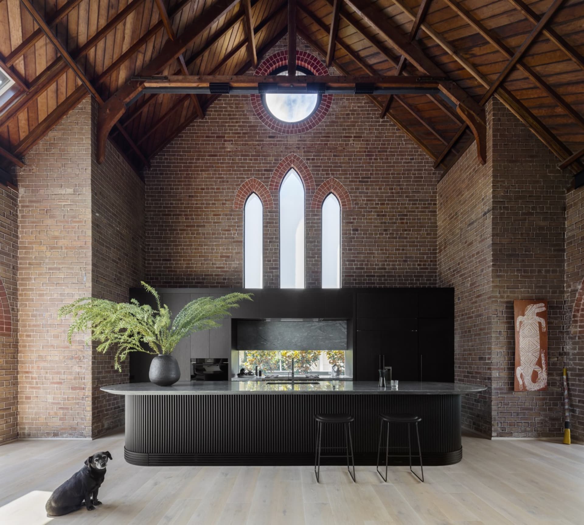 A modern farmhouse kitchen with a brick wall and concrete countertops featuring a white oak floor, black island, and black ba