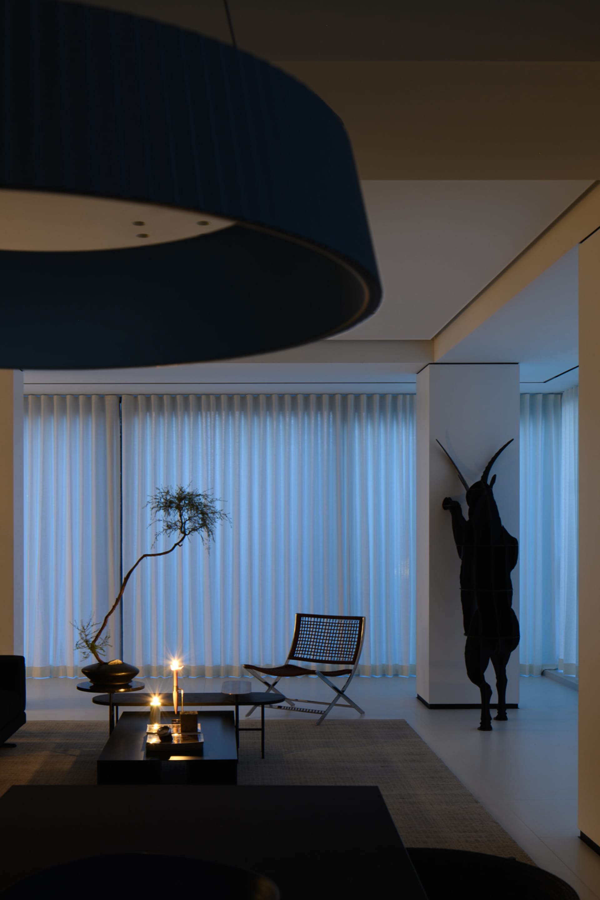 A minimalist living room interior with white curtains, a glass table, metal chairs, and a black pendant light.