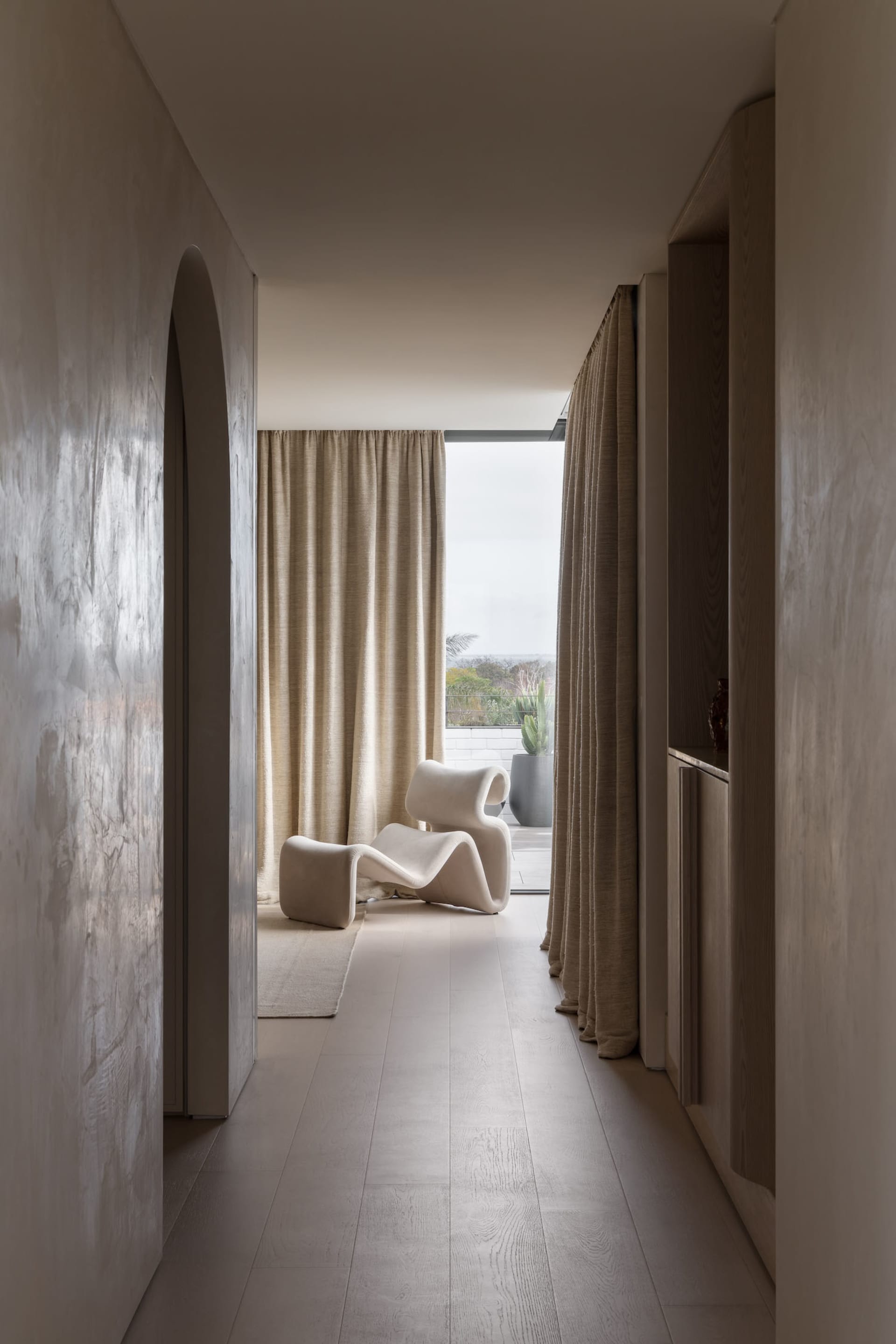 A minimalist living room with modern furniture and neutral tones, featuring a white chair and a view of the outdoors.