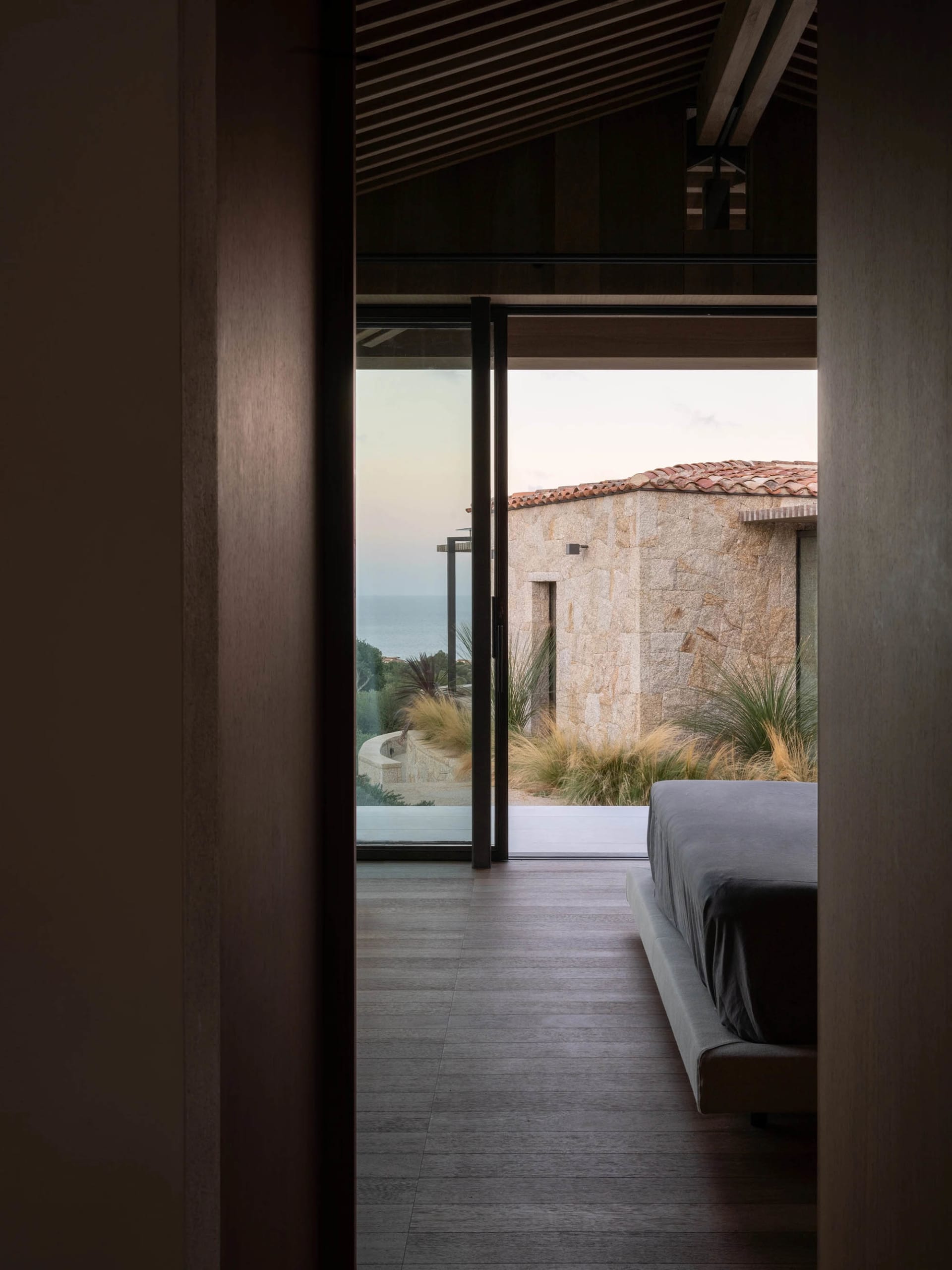 A modern bedroom with wooden flooring, a gray bed, and a view of a house with a red-tiled roof and sea in the background.
