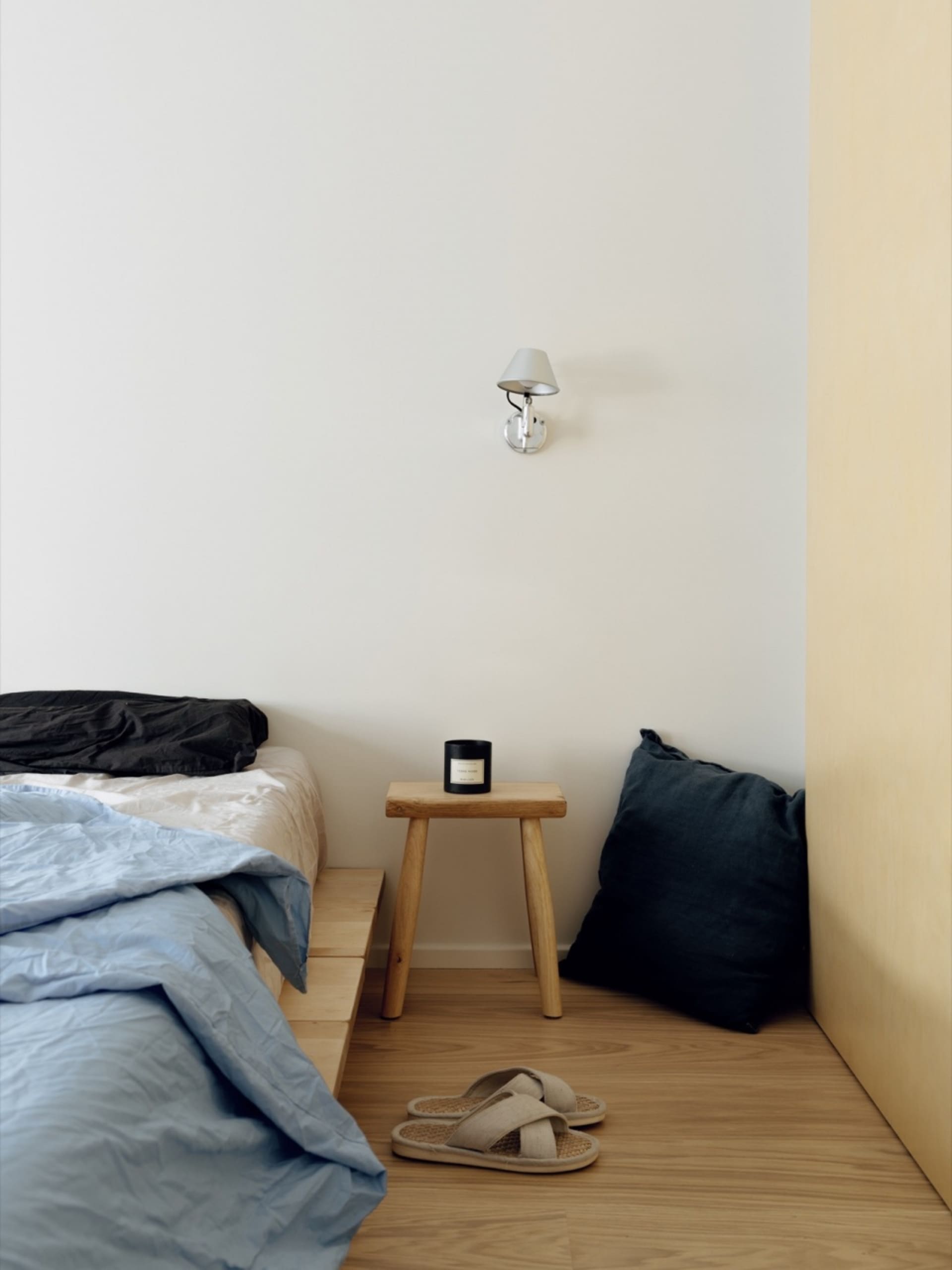 Minimalist bedroom with a bed, stool, and slippers on a wooden floor, featuring a wall-mounted light fixture.