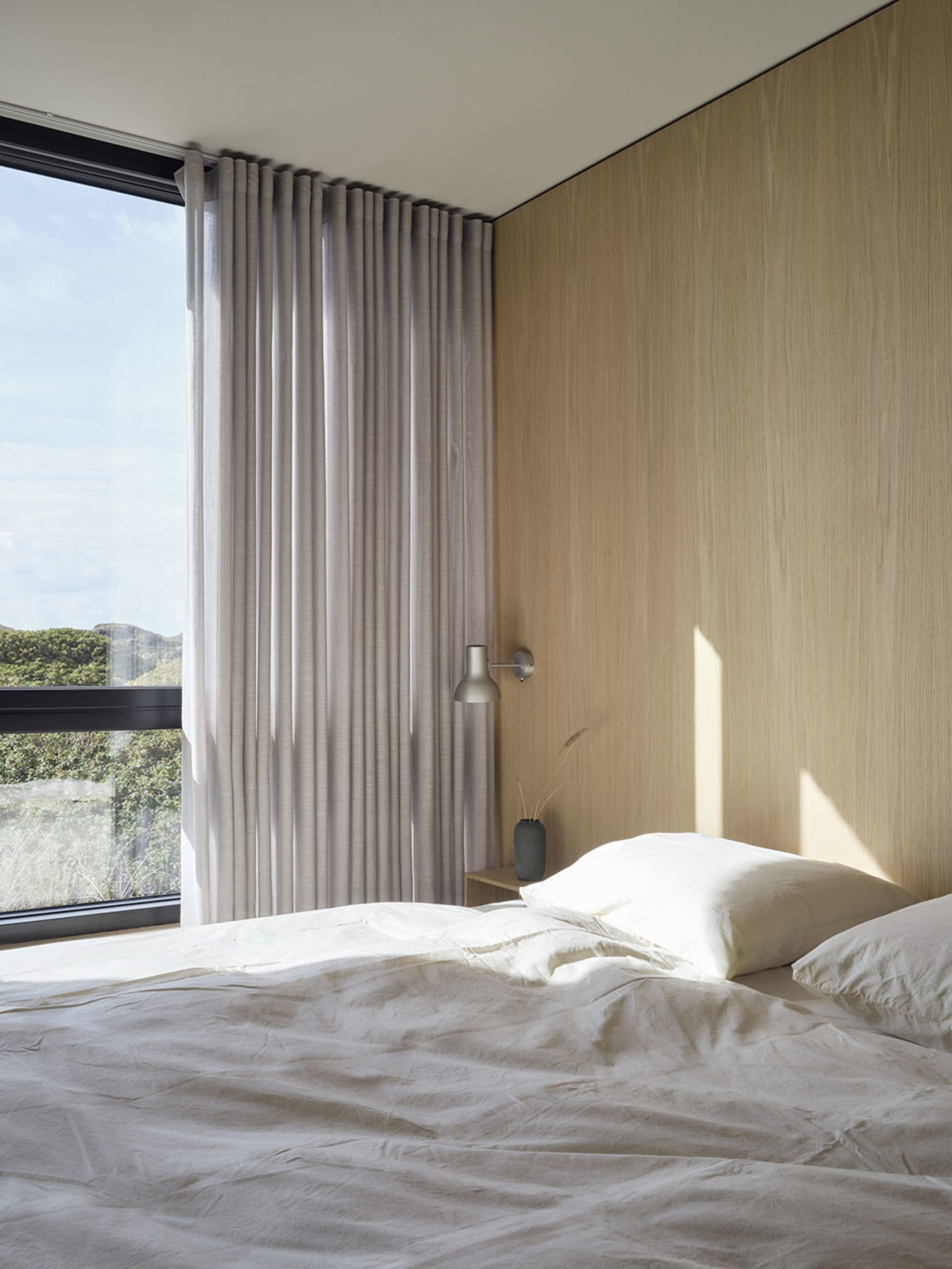 A minimalist bedroom with a textured wooden wall and a grey bed against a large window.