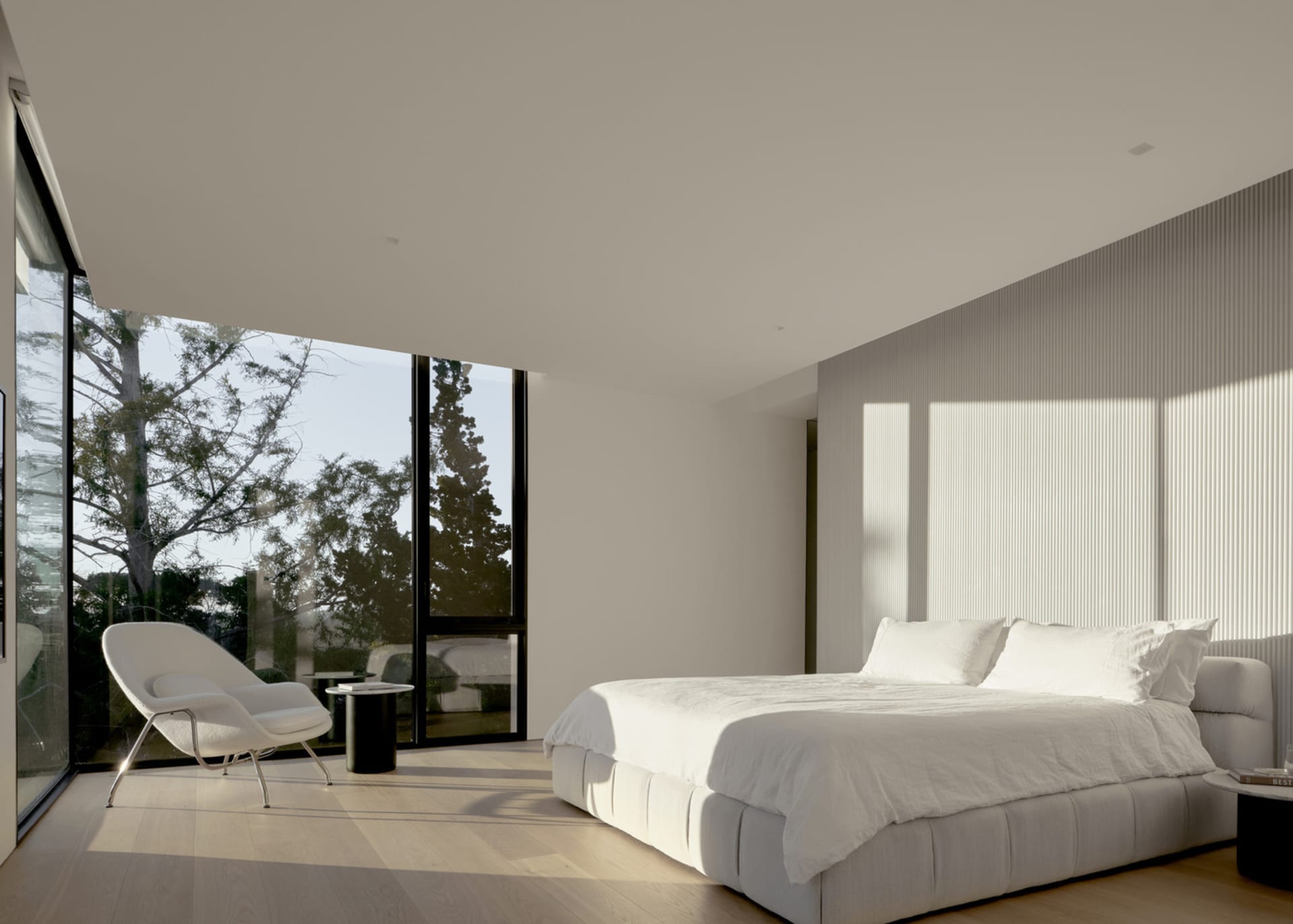 A minimalist bedroom featuring a white bed with a sleek, modern chair in front of a large bay window with a view of trees.
