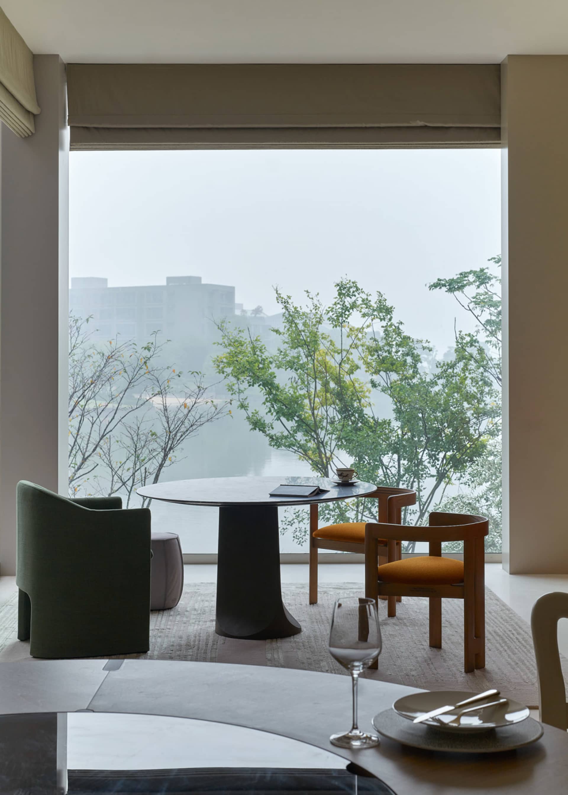 A modern restaurant with a round table and chairs next to large windows overlooking water and trees.