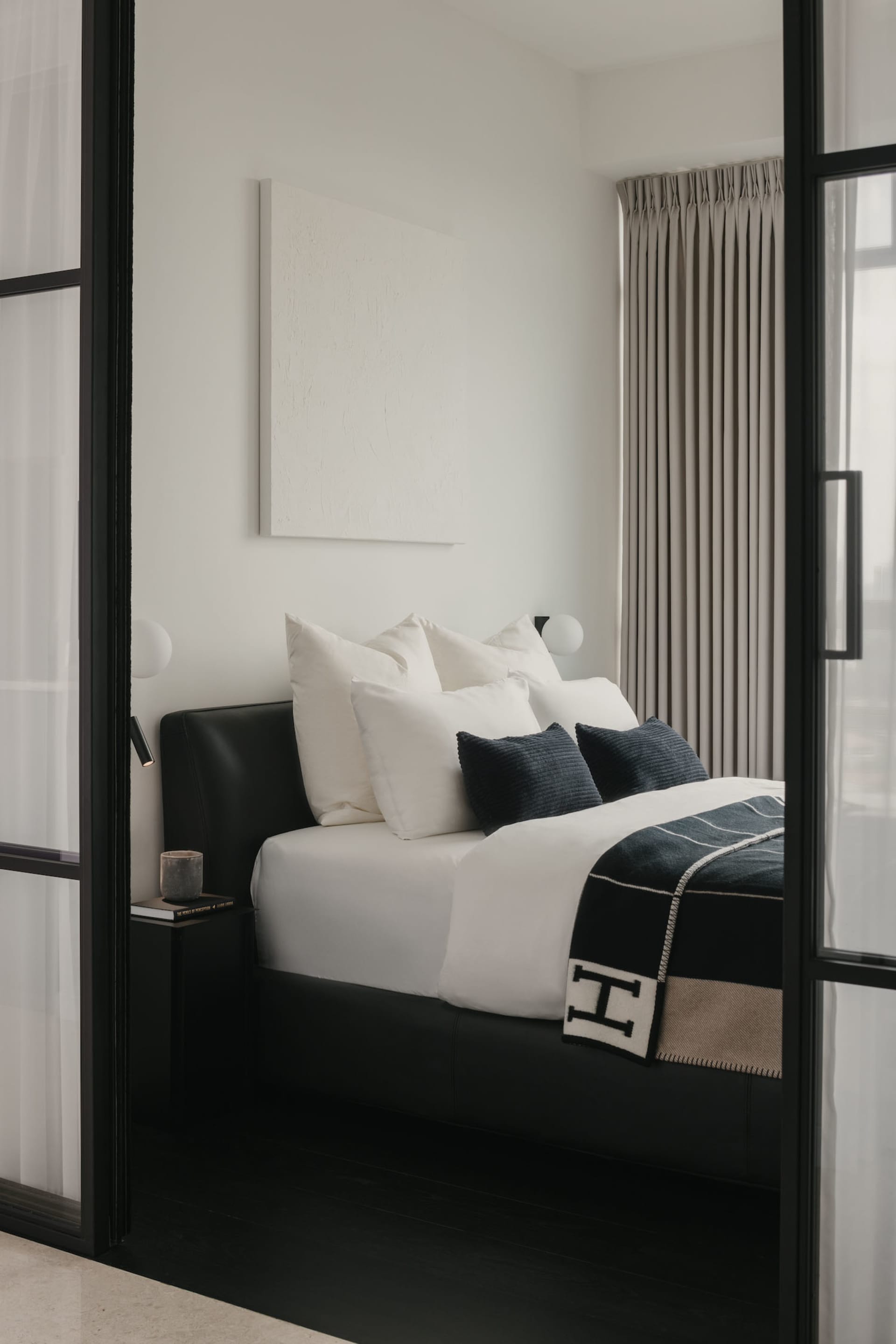 Minimalist bedroom featuring a bed adorned with black and white textiles, positioned near tall, closed drapes.