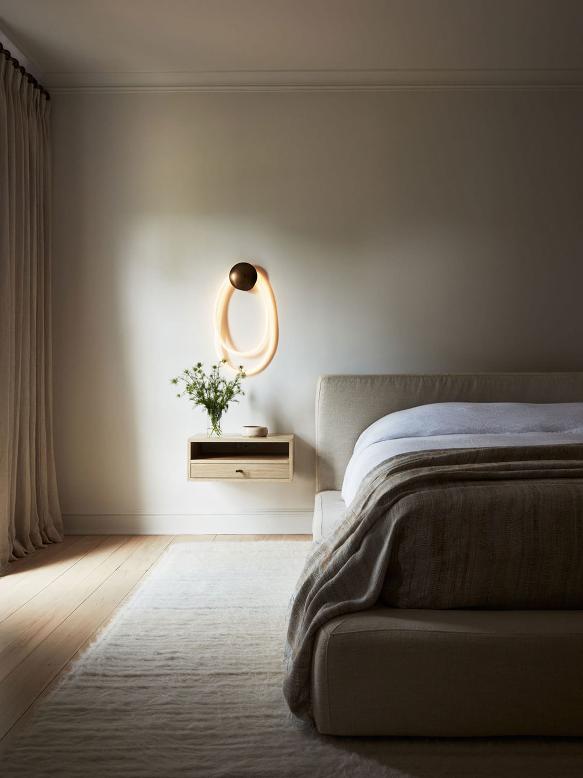 Minimalist bedroom with fabric bedding on a wooden bed frame, featuring a floating nightstand and a modern light fixture.