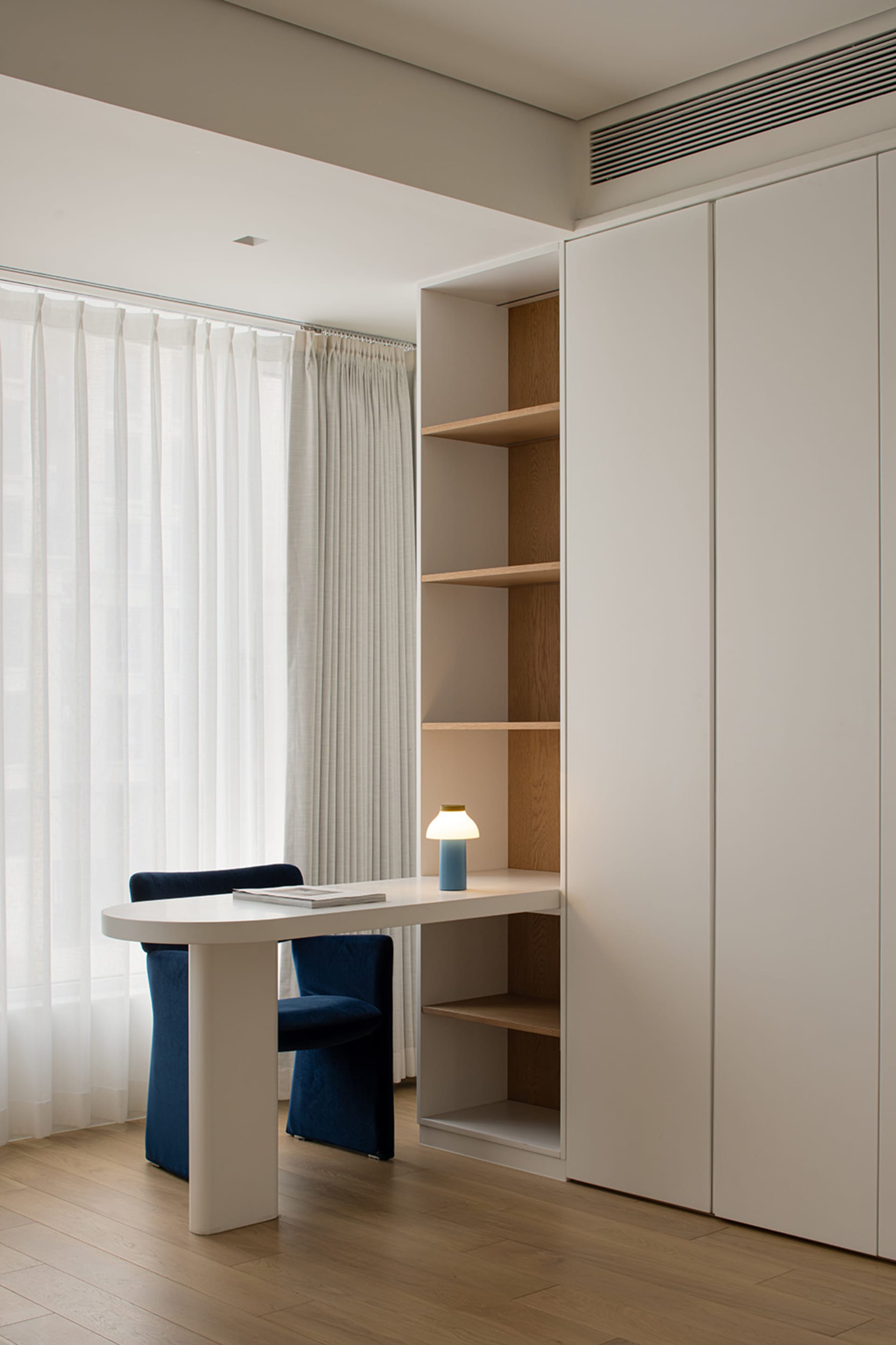 A minimalist home office featuring a white desk, blue chair, and white oak shelves.