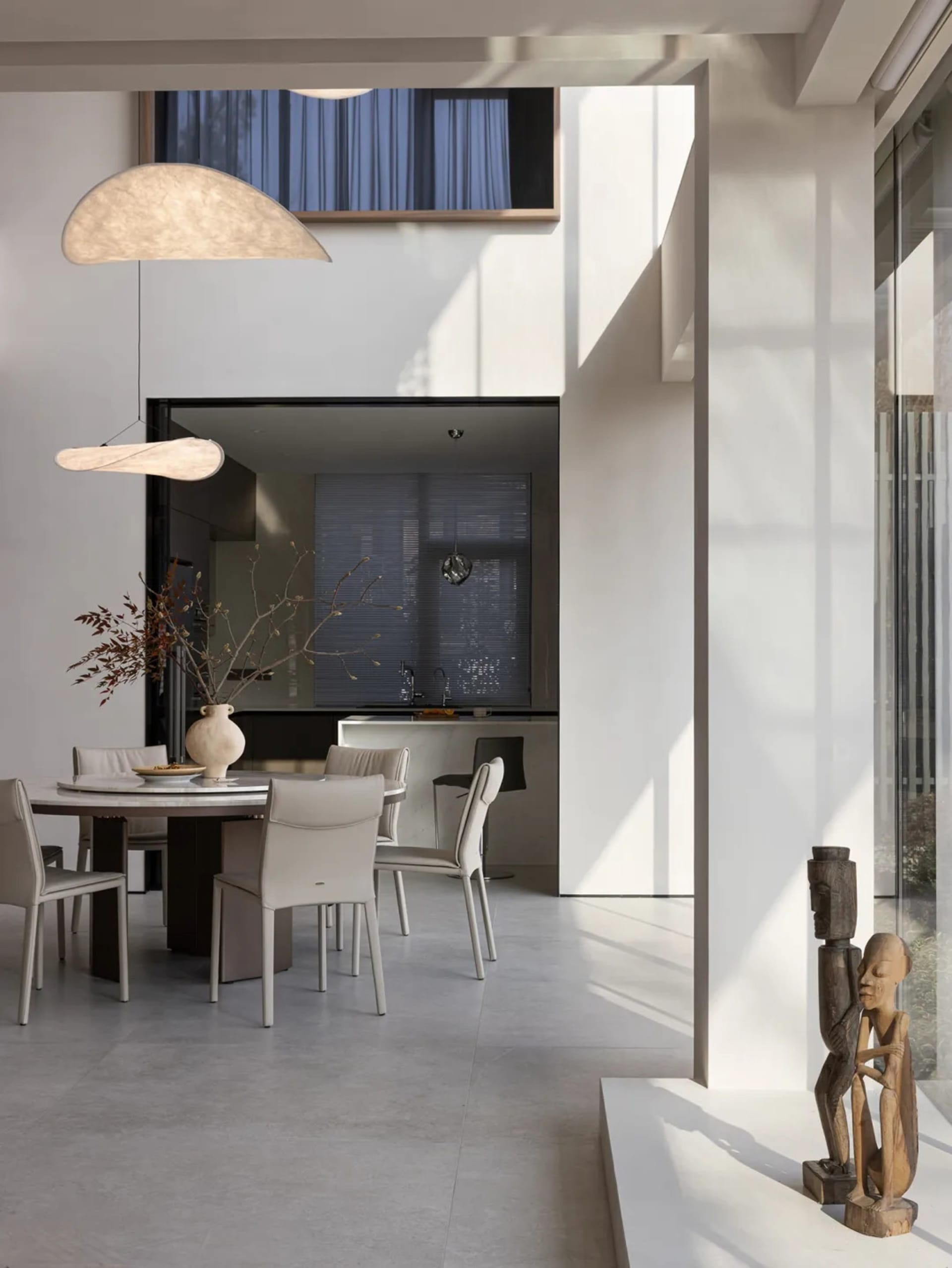 Modern dining room with a round table, white chairs, and a vase of dried flowers on the table, with a kitchen area in the bac