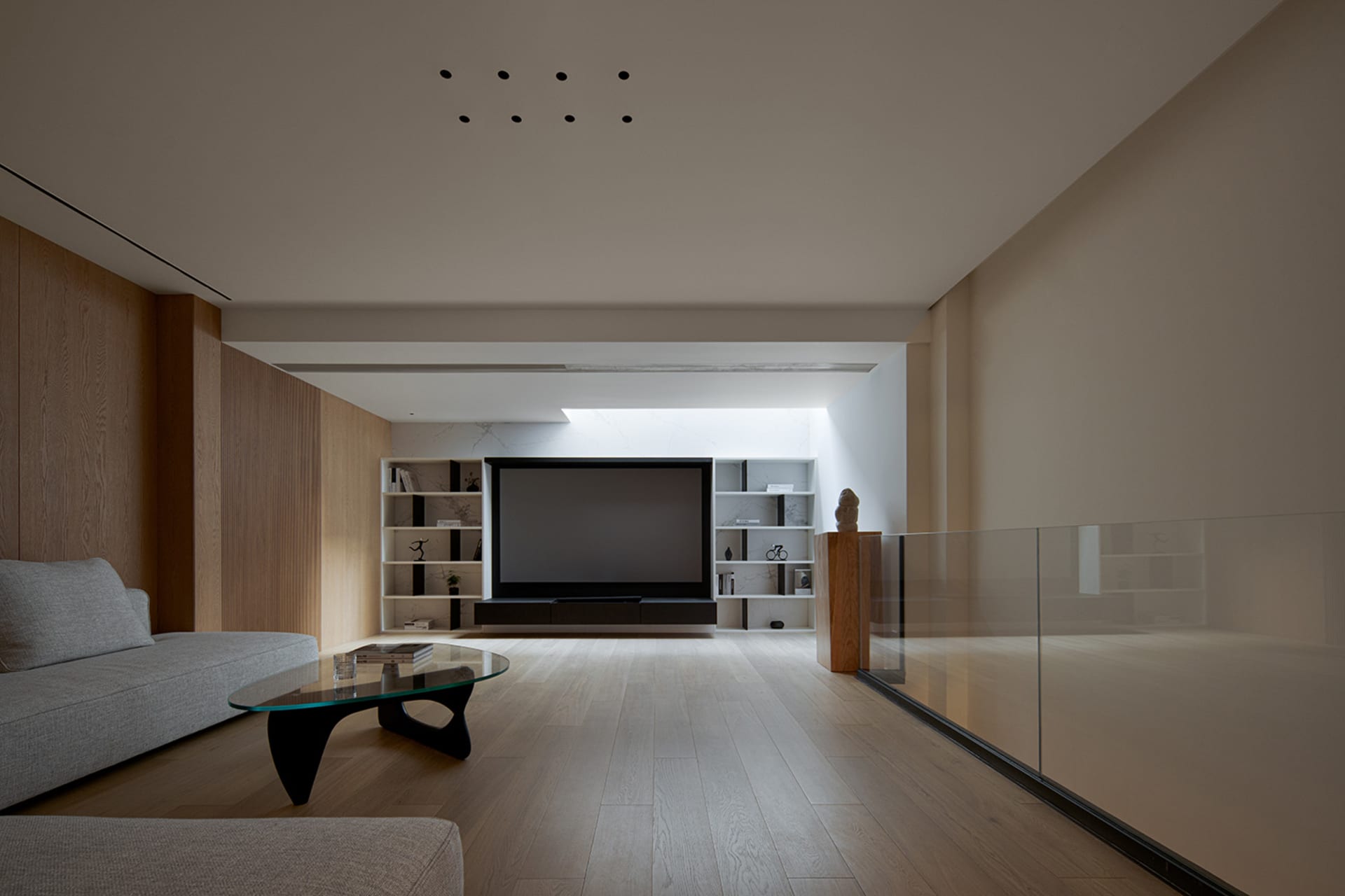 A modern living room featuring a minimalist design with wood flooring, a glass coffee table, and a large screen TV.