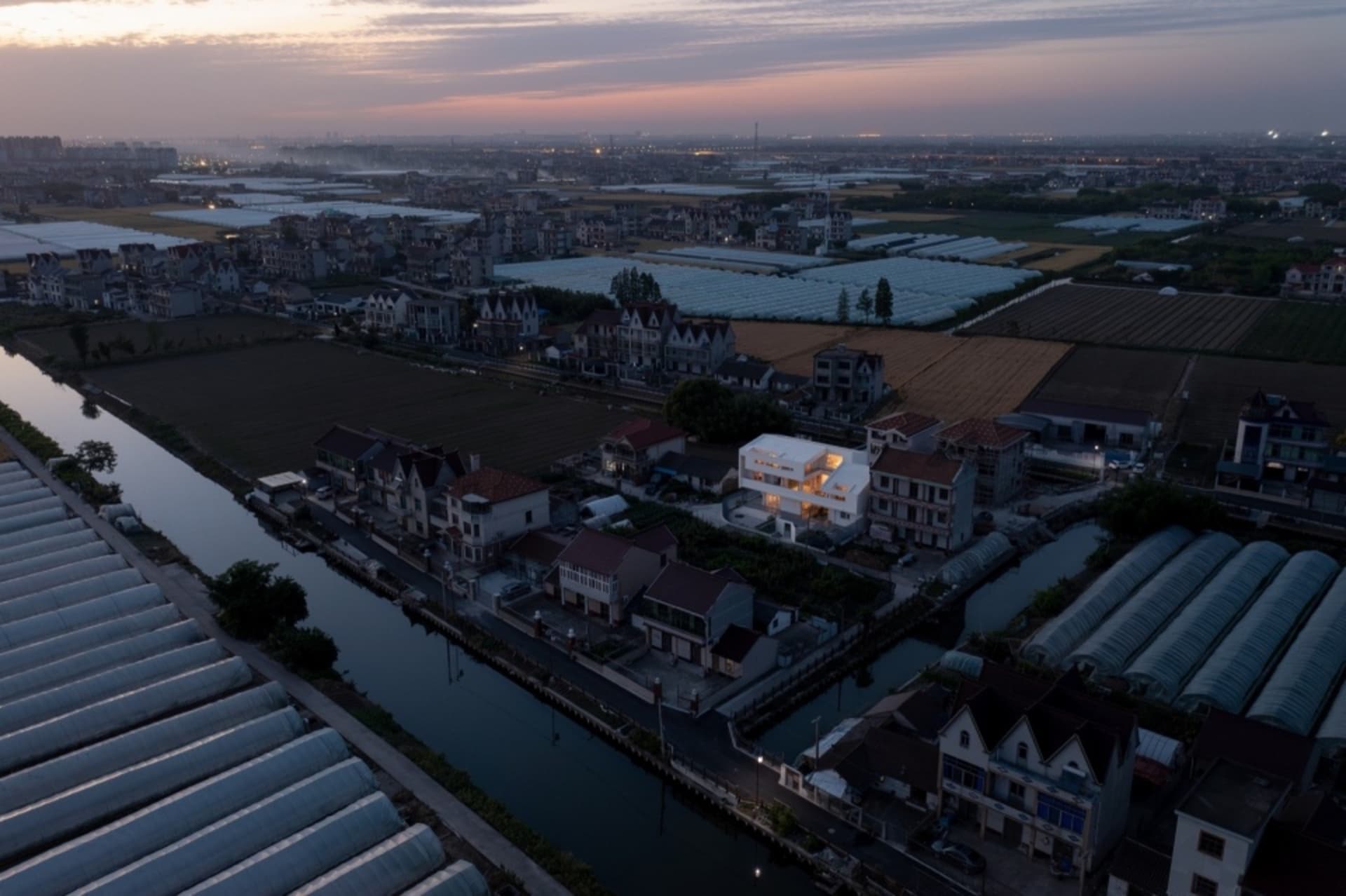 A village with greenhouses and agricultural fields during dusk with a sky gradient from purple to pink.