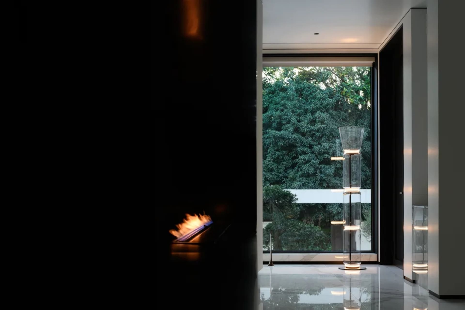 A modern entryway featuring a glass fireplace, large windows overlooking lush greenery, and sleek floor lamps.