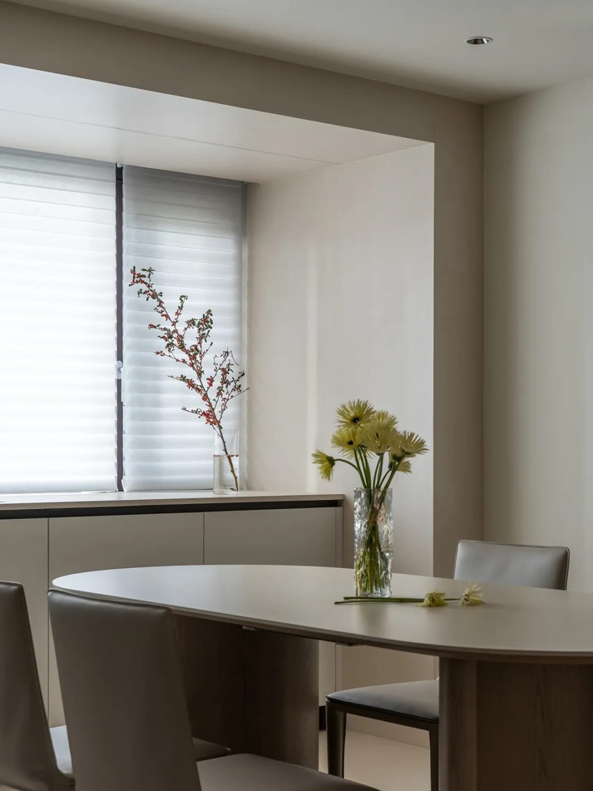 Modern dining room with minimalist furniture and decor, including a round table, chairs, and fresh flowers in a vase by the w