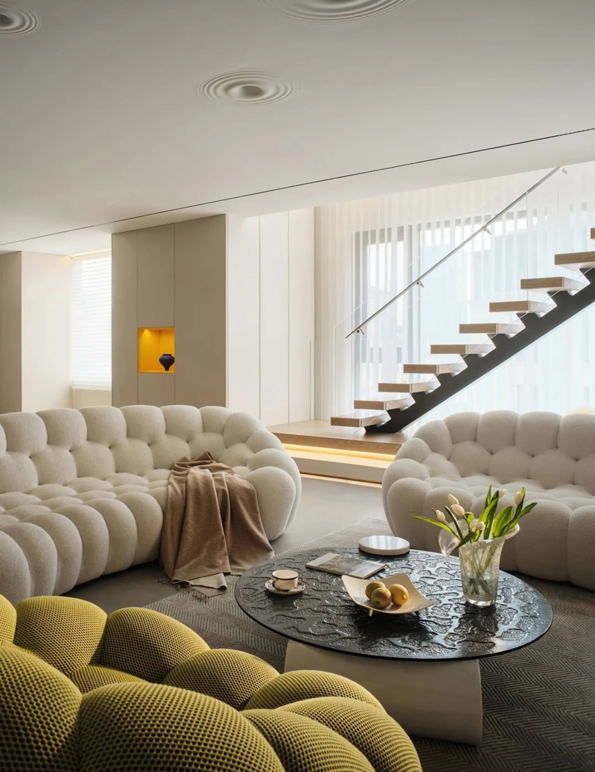 Modern living room featuring plush white sofas with yellow accents, a black coffee table, and a staircase in the background.