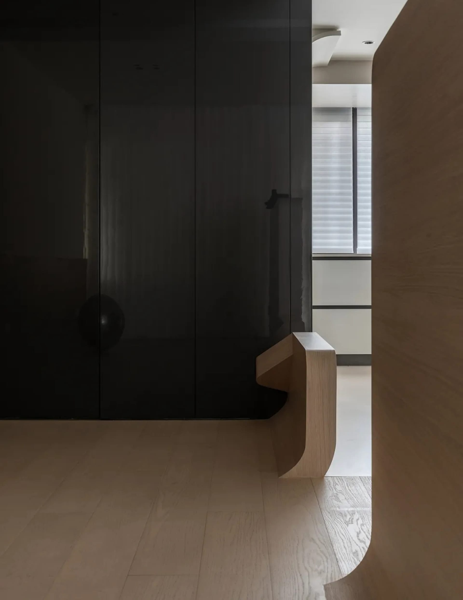 A minimalist entryway with a black glass wall and a wooden bench on white flooring