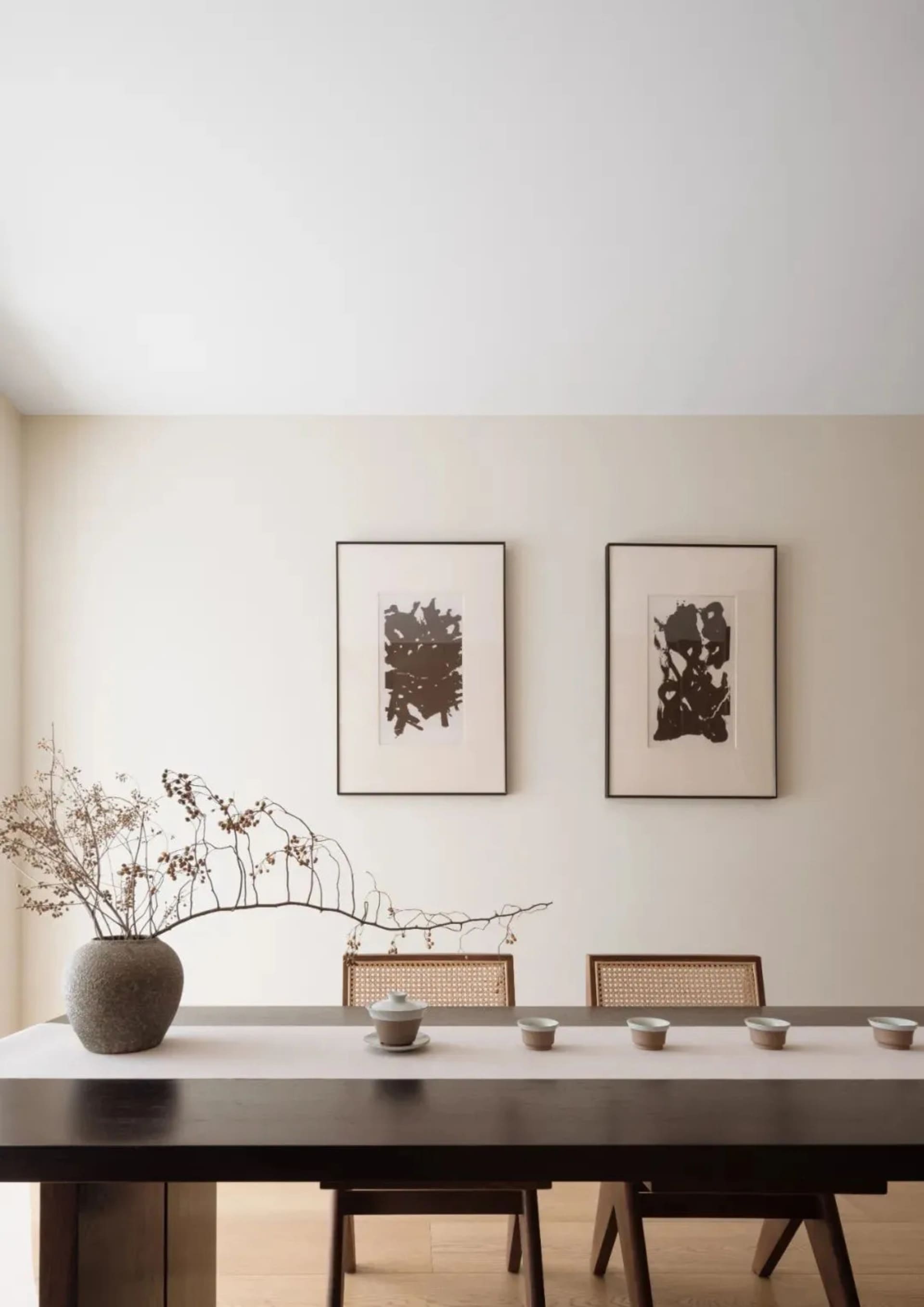 Modern dining room with wooden table and cane-back chairs, minimalistic art on the wall, and a vase with dried branches.
