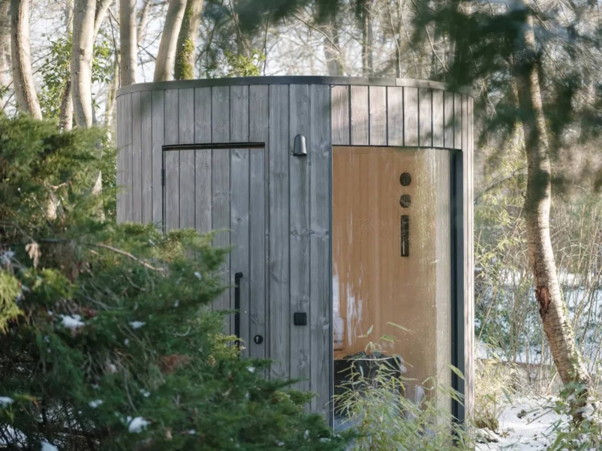 A modern outdoor spa with a wooden exterior surrounded by trees and snow-covered ground.