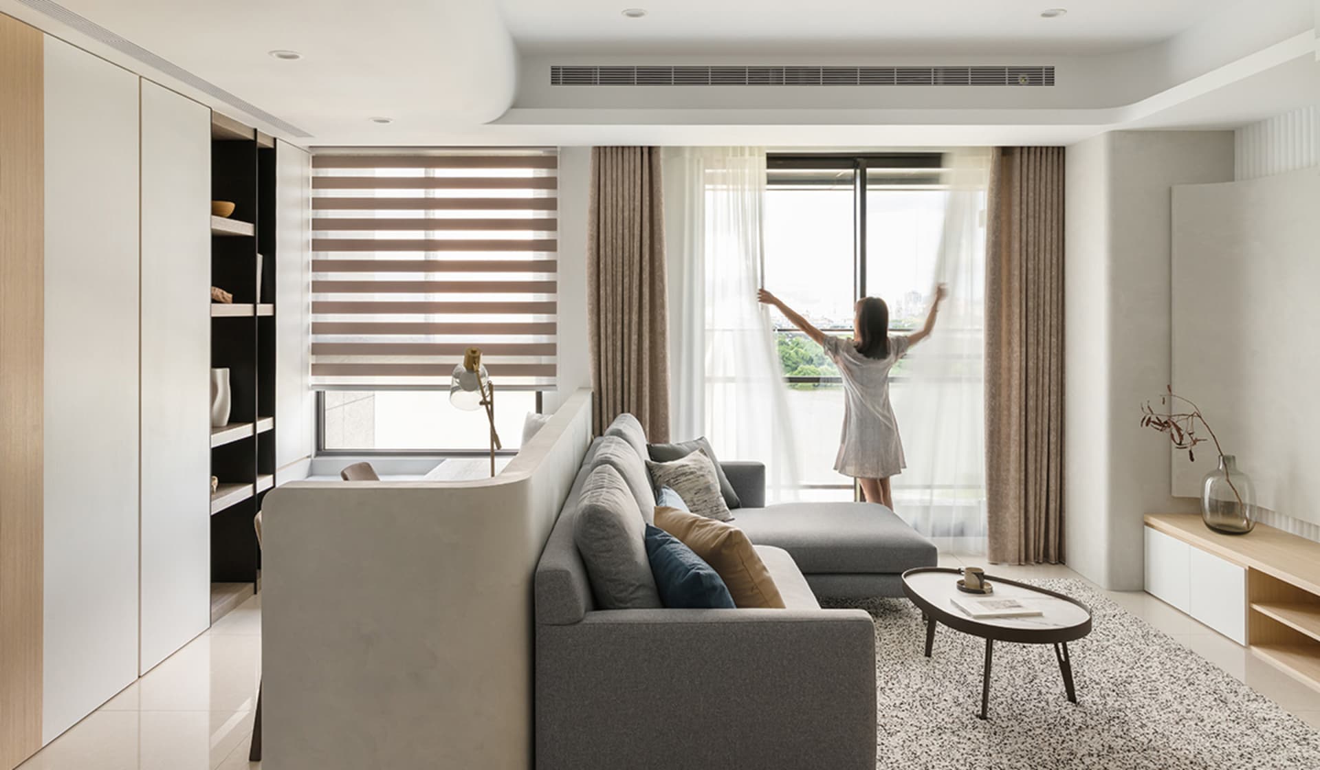 Person adjusting beige curtains in a minimalist living room with a gray sectional sofa and a round coffee table.