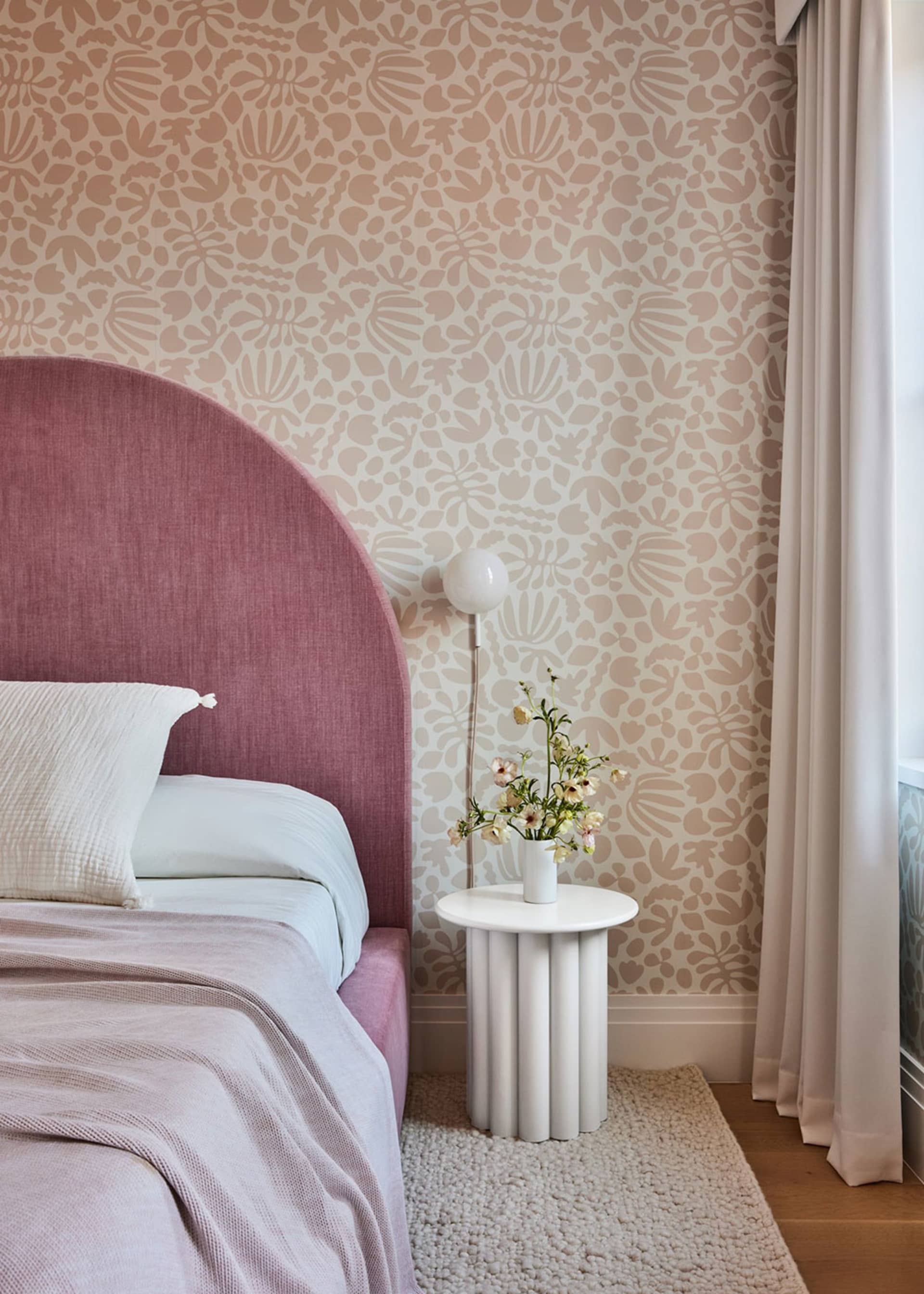 A modern bedroom featuring a bed with a pink upholstered headboard, a white side table with a vase of flowers, and patterned
