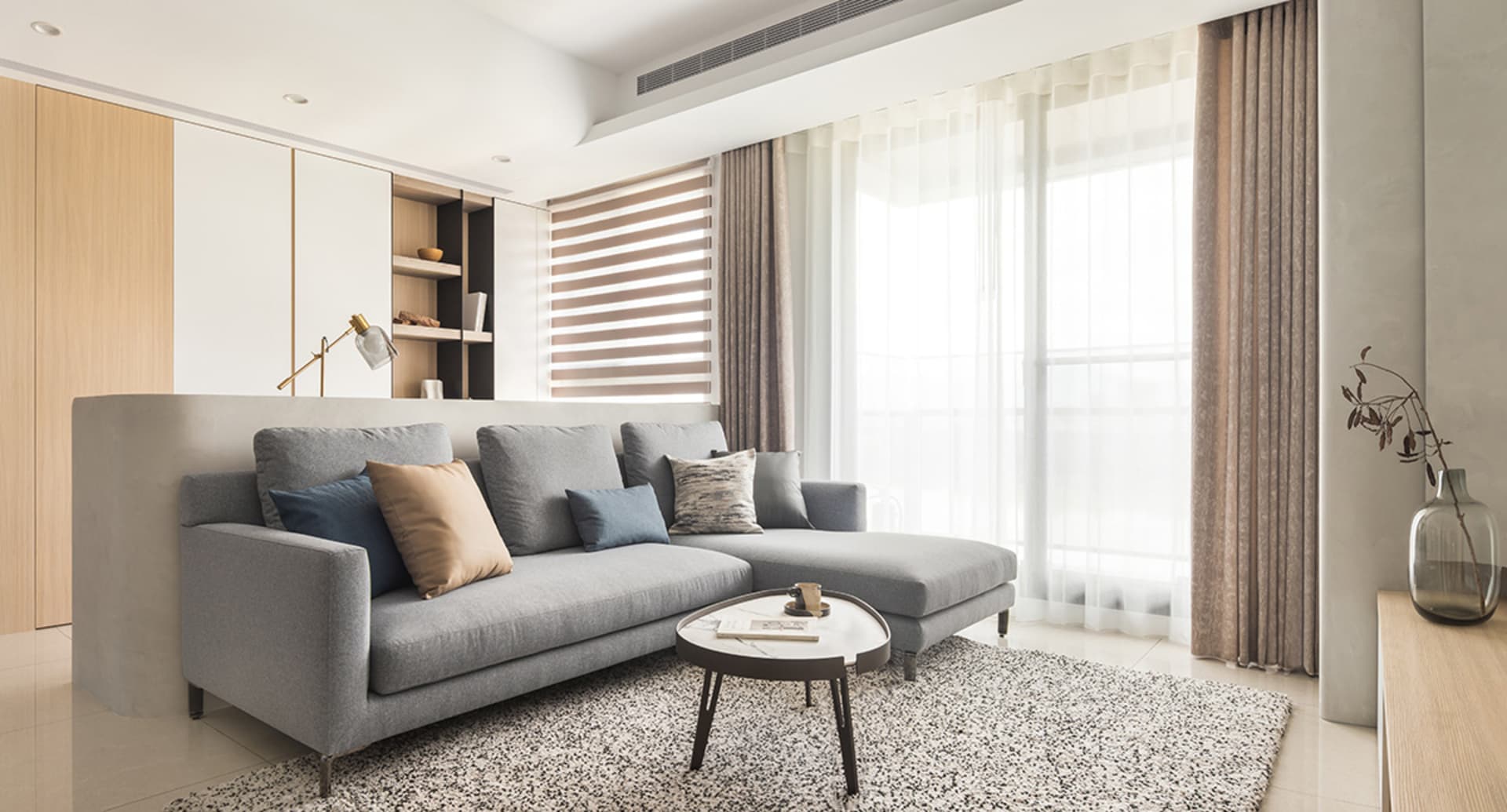 A modern minimalist living room with a gray sectional sofa, beige cushions, and a round coffee table on a patterned rug.