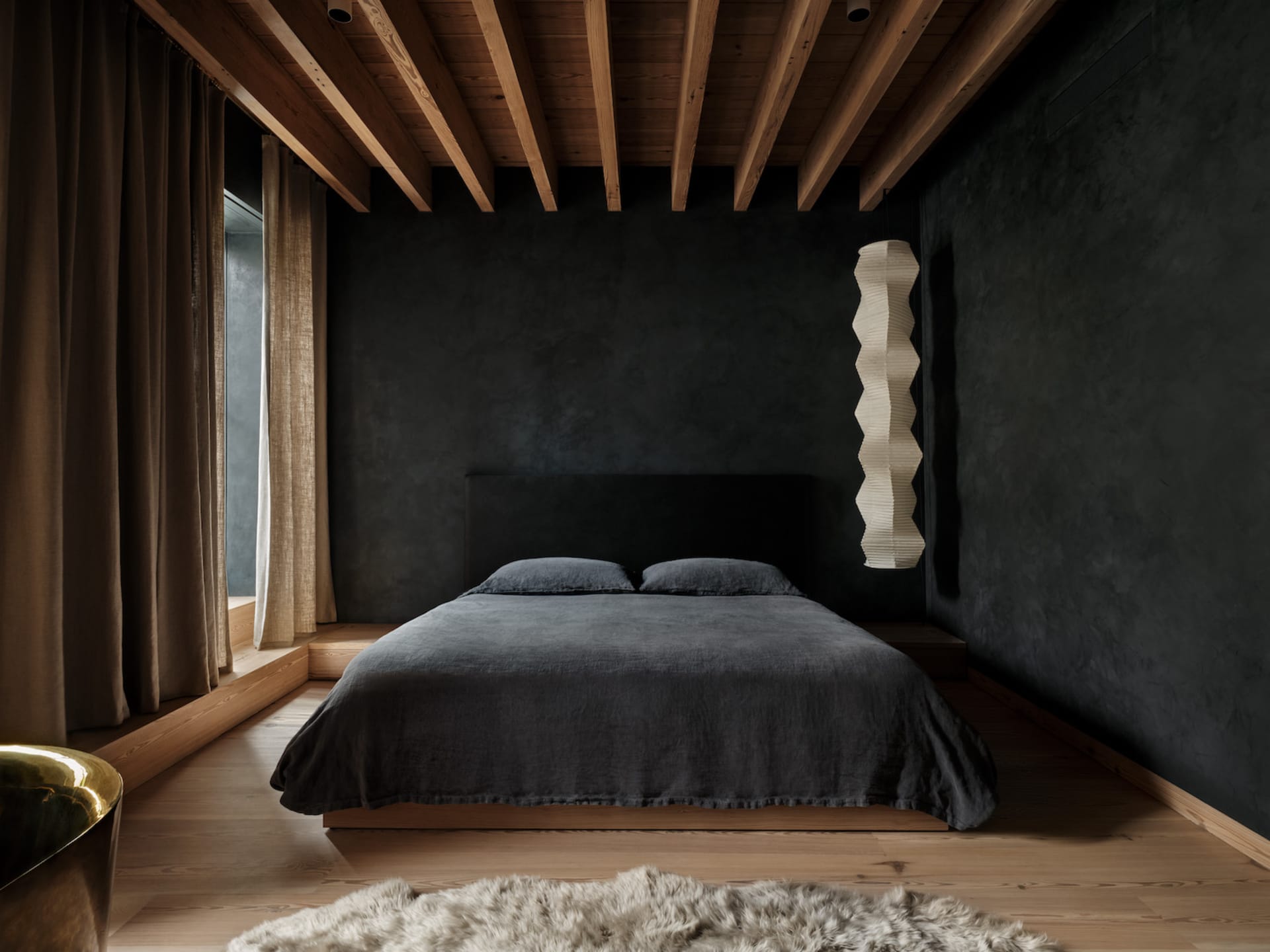 A minimalist bedroom with dark walls and wood flooring, featuring a low platform bed with a dark headboard and bedding, and a