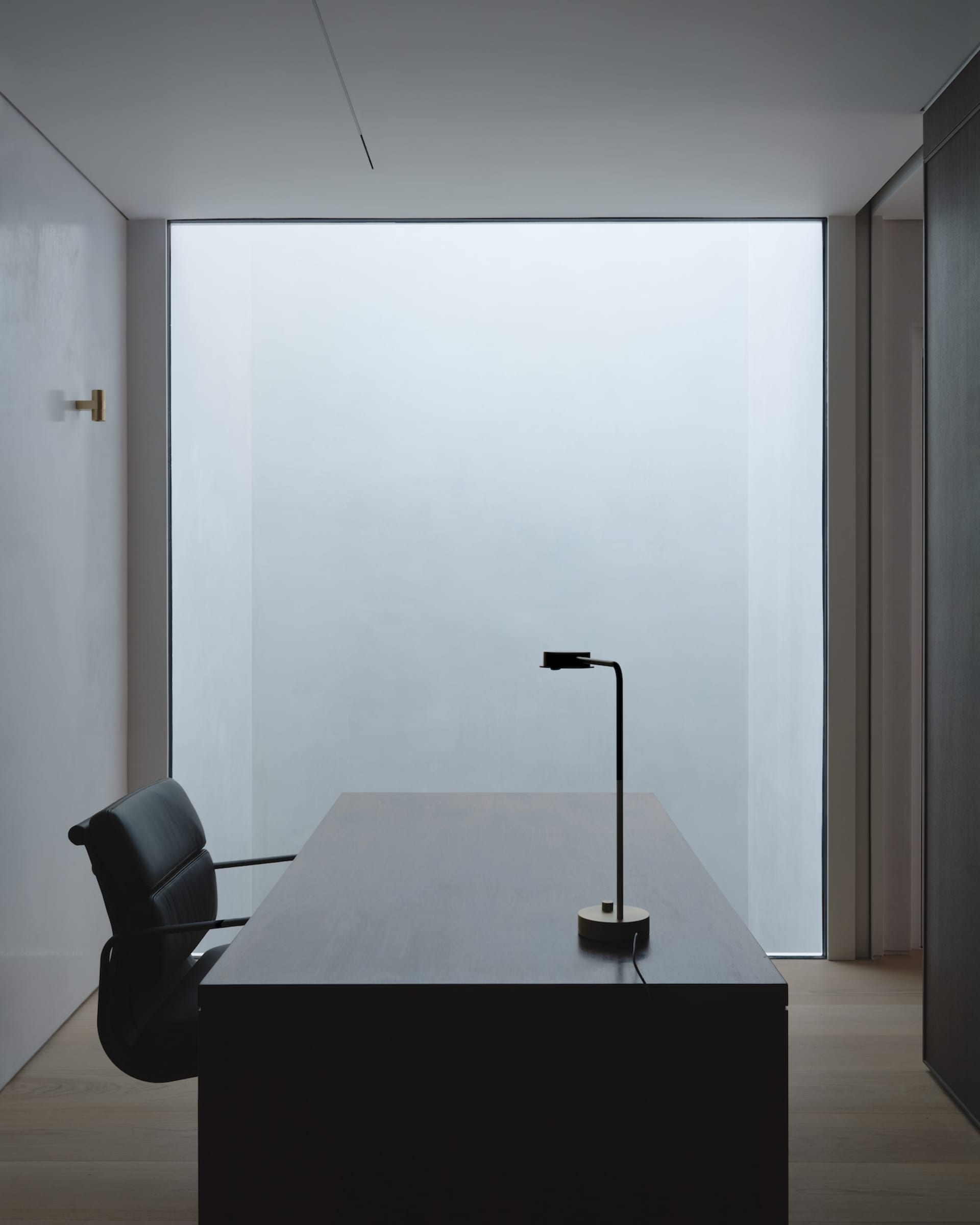 Minimalist office space featuring wooden desk and glass window with black chair.