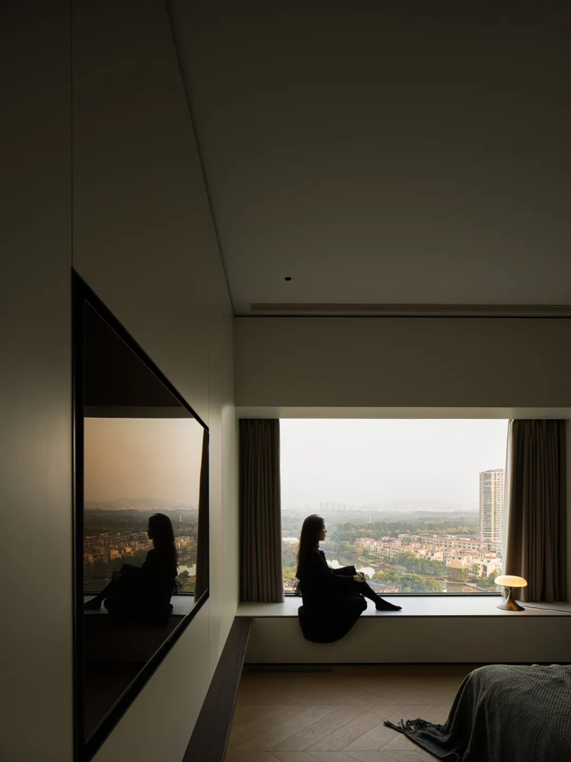 A person sitting on a window sill in a modern interior with glass walls, wooden flooring, and textile furnishings.