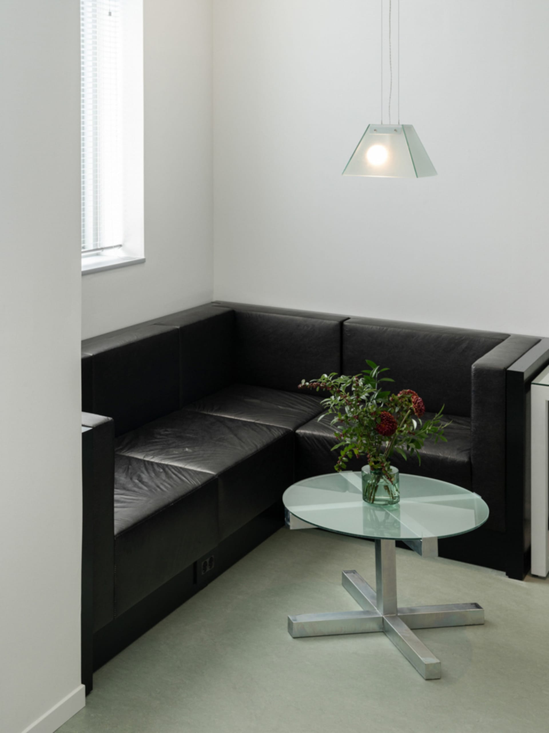 A minimalist living room featuring a black leather sofa, a round glass coffee table, and a hanging pendant light.