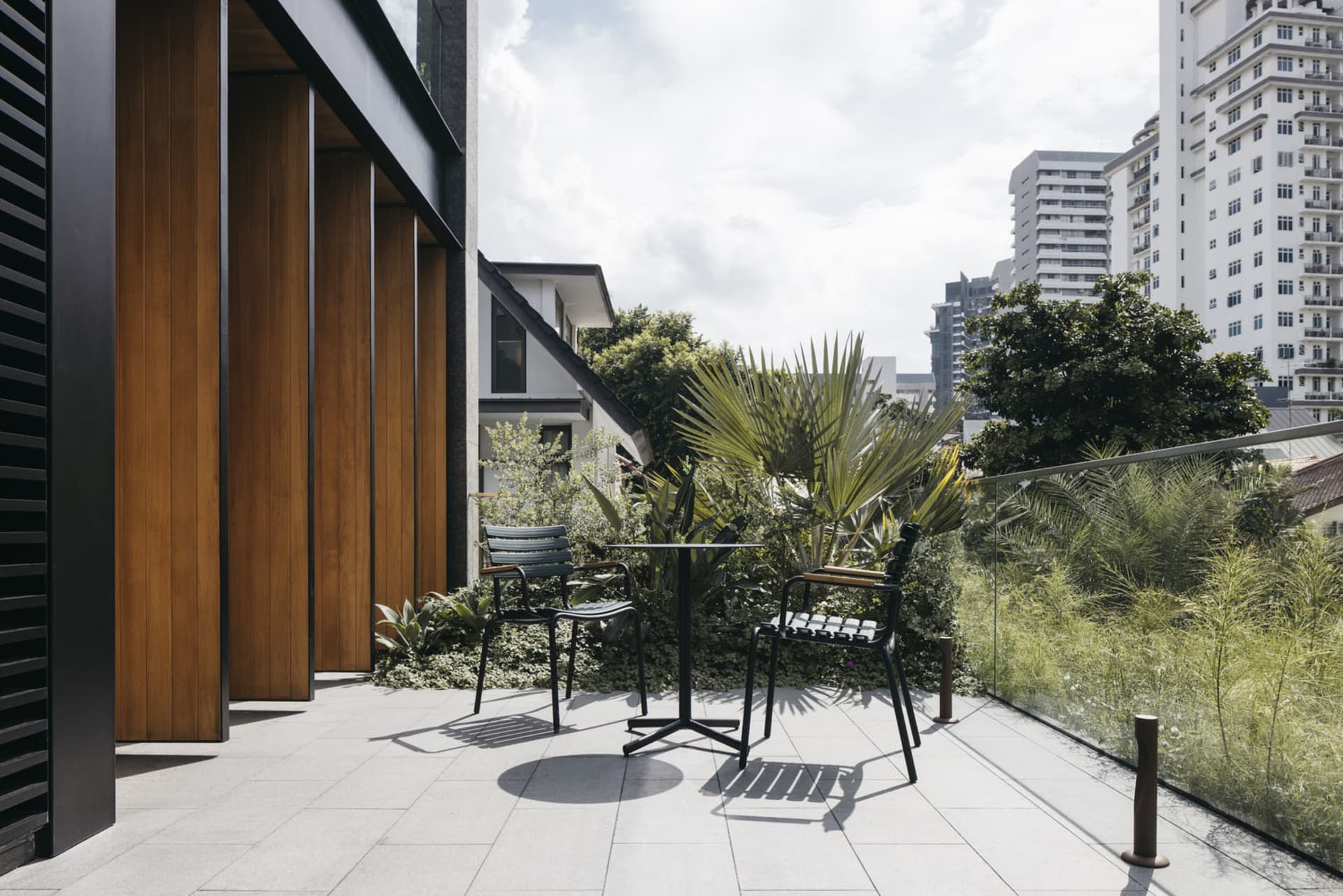 Concrete patio with modern black metal chairs and table, surrounded by greenery and topped with tropical plants.