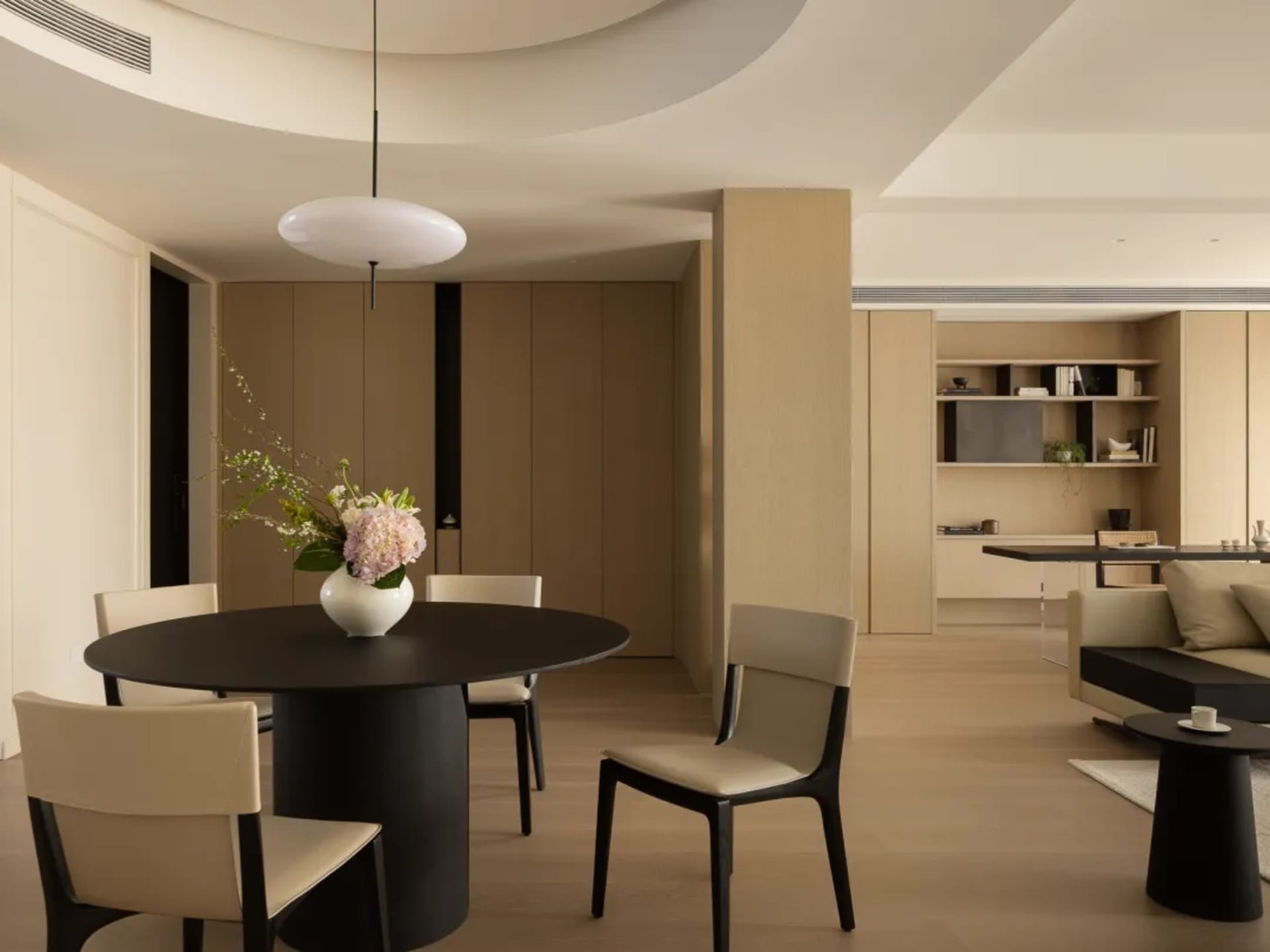 Modern living room with wooden board floor, beige furniture, and a black dining table with a vase of flowers.